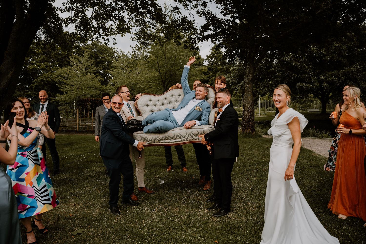 Happy wedding guests gather outdoors while playfully tossing a person in the air during reception celebration.