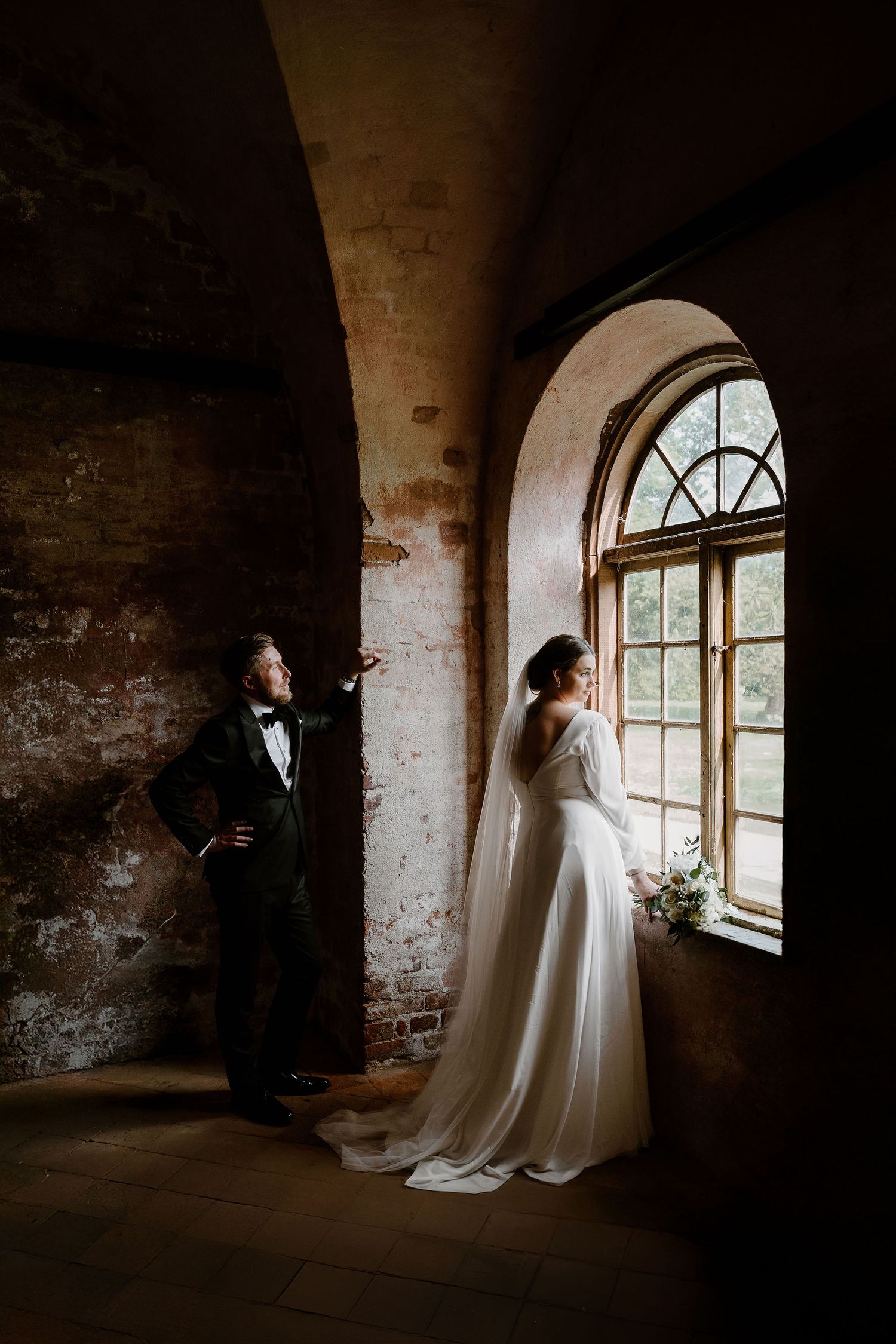 A wedding couple poses separately in an atmospheric stone room with arched windows and weathered brick walls.