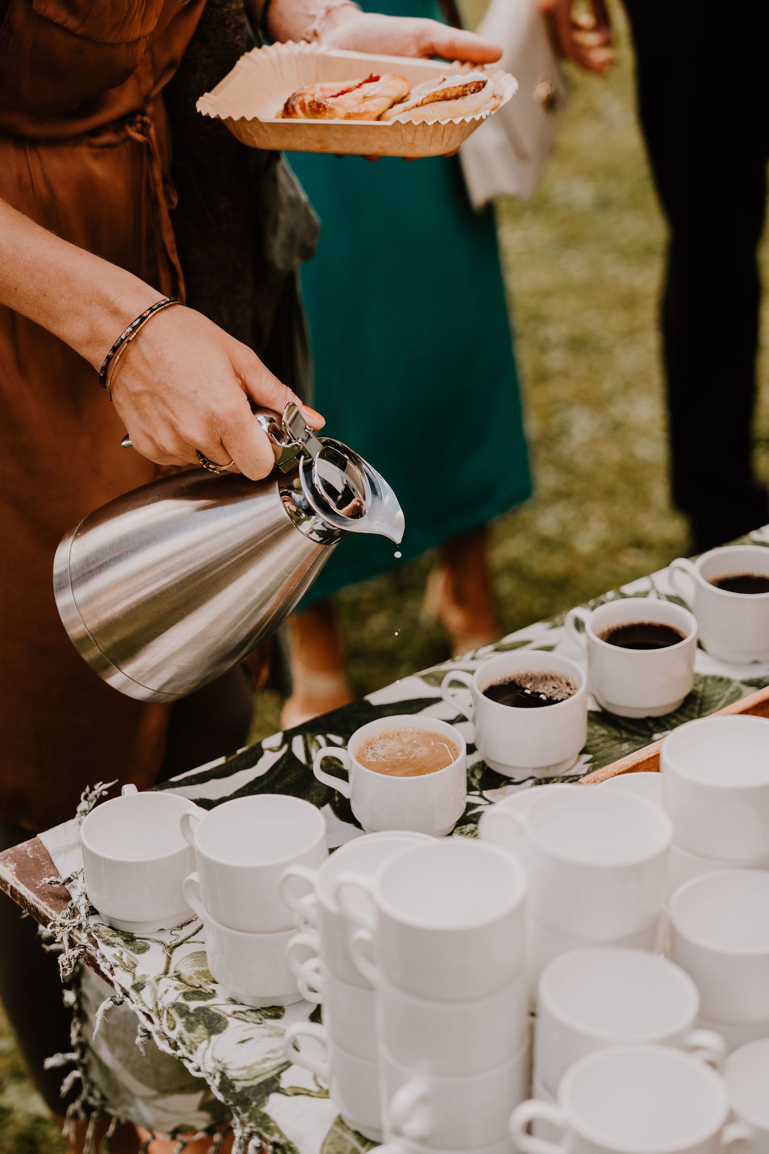 Someone pours coffee from a silver carafe into white cups at an outdoor gathering while holding a plate of desserts.
