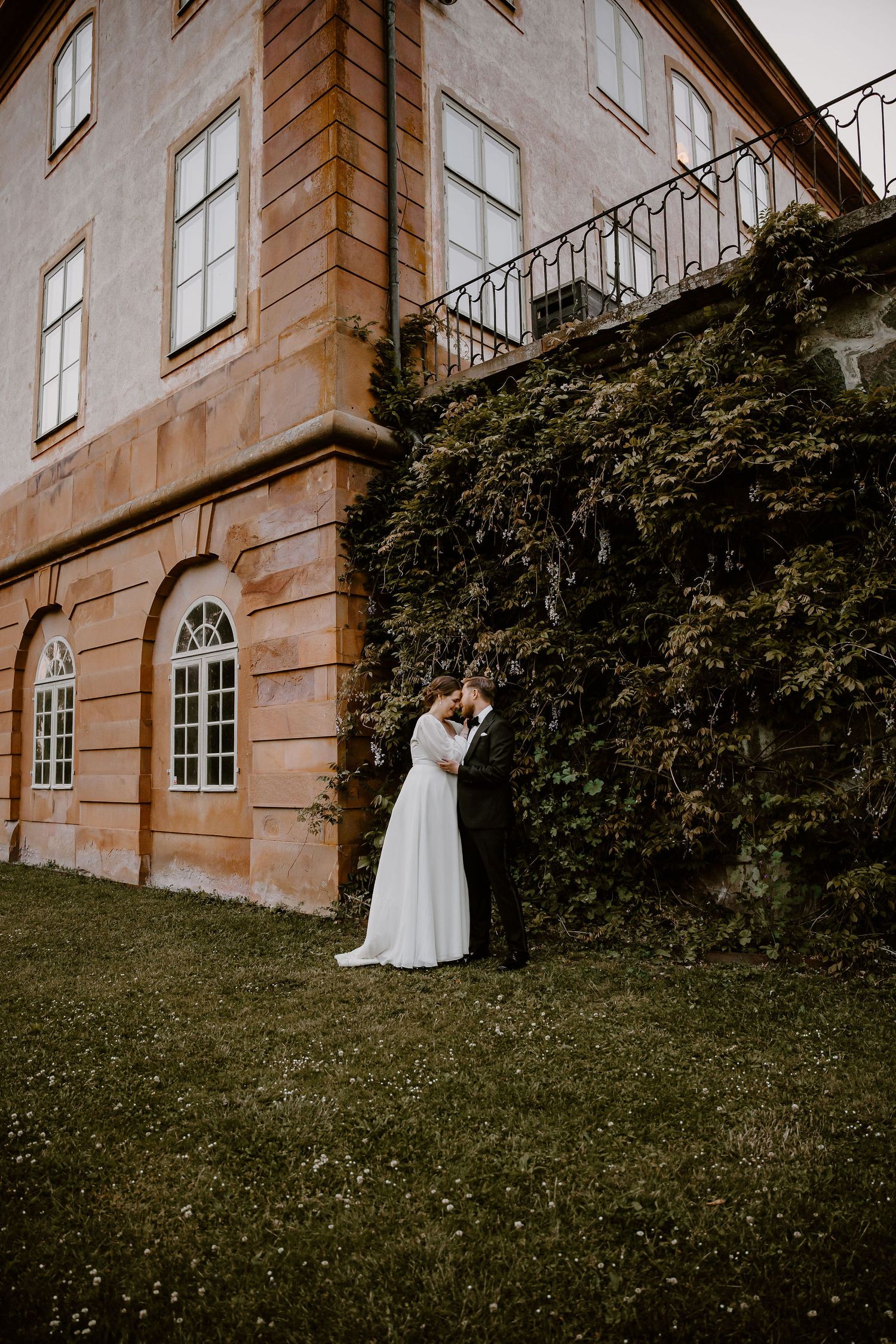 Elegant couple shares intimate moment on grassy hillside against backdrop of historic stone mansion at golden hour.