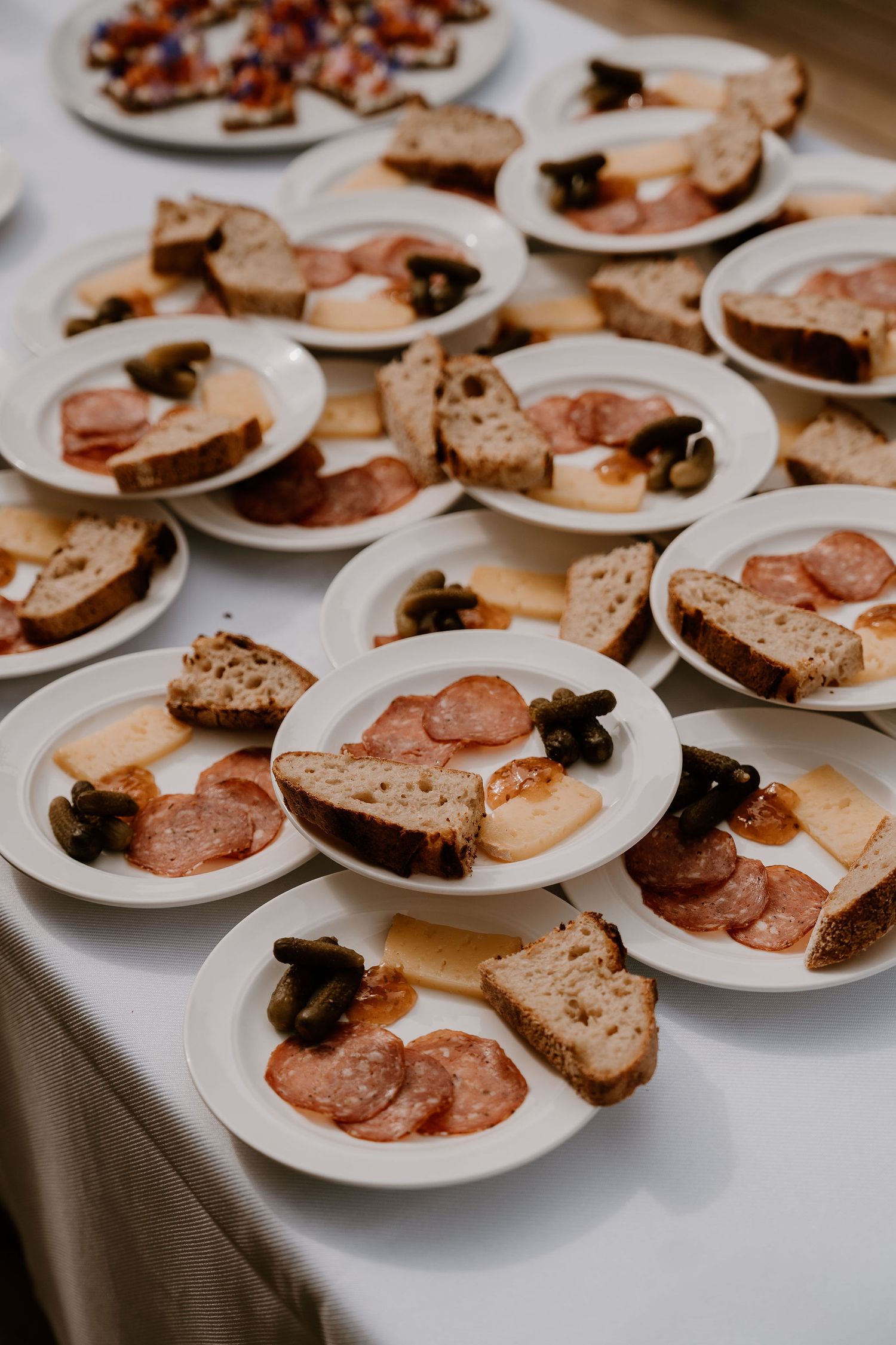Multiple plates of sliced bread and cured meats arranged on a white tablecloth at a catering event.