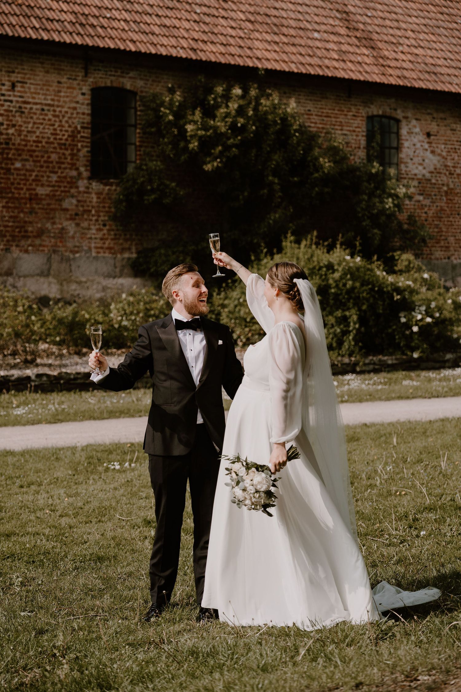 A bride and groom toast with champagne glasses outdoors in front of a rustic brick building on their wedding day.