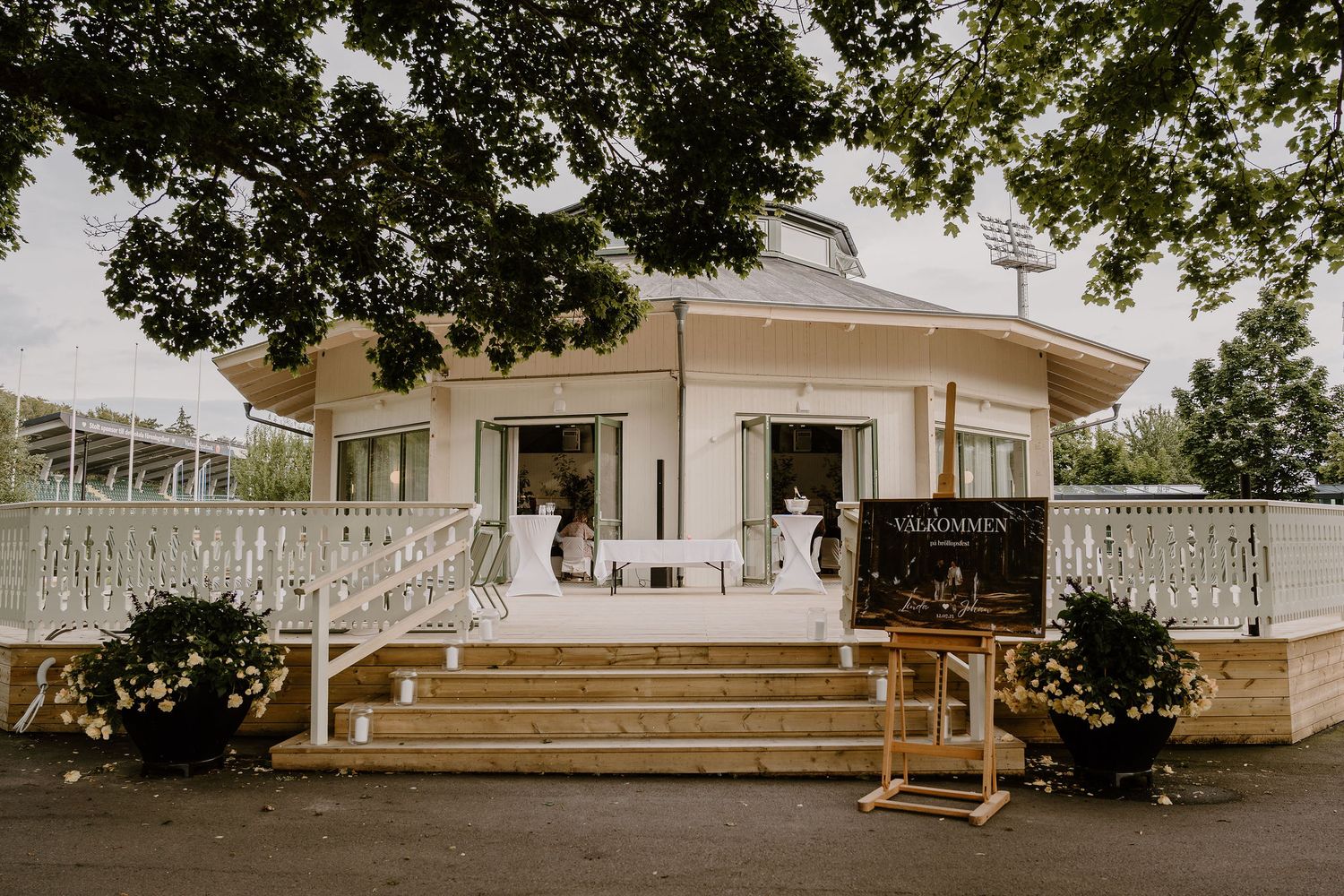 A scenic country wedding venue with white buildings and rustic wooden benches set beneath mature shade trees.