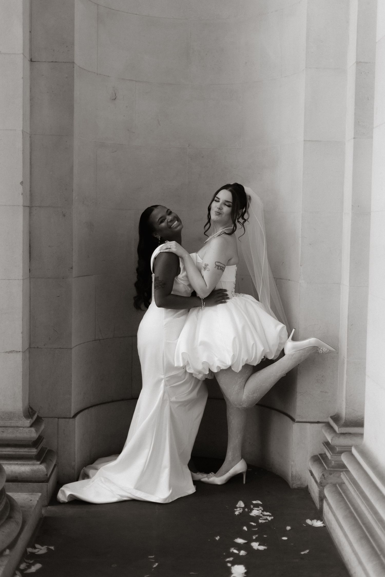 Black and white photo of two brides in white gowns posing playfully against stone columns.
