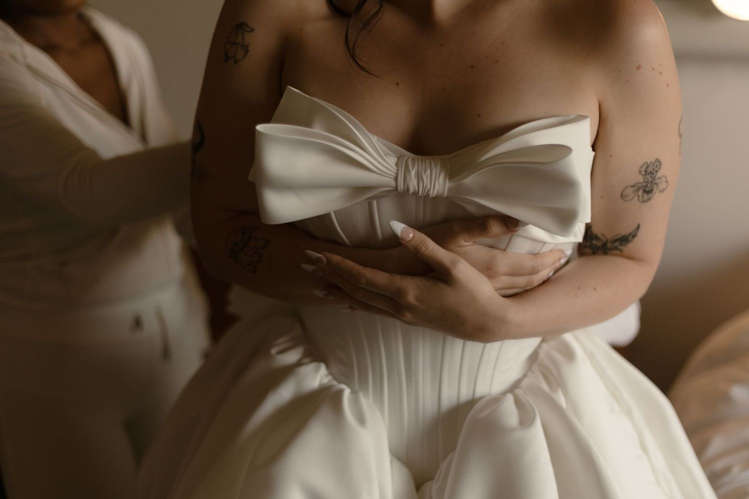 Bride in strapless white wedding gown with large bow detail, assisted by tattooed bridesmaid during bridal preparation.