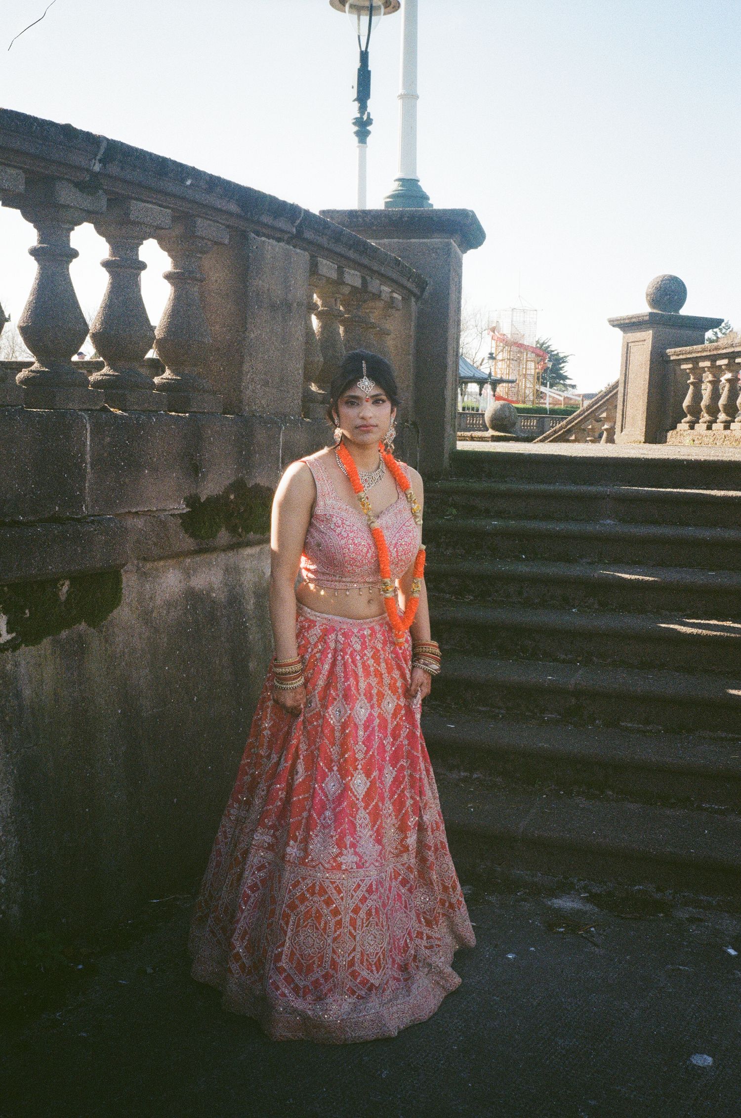 Woman in ornate pink and orange traditional Indian lehenga poses on stone steps at outdoor location.