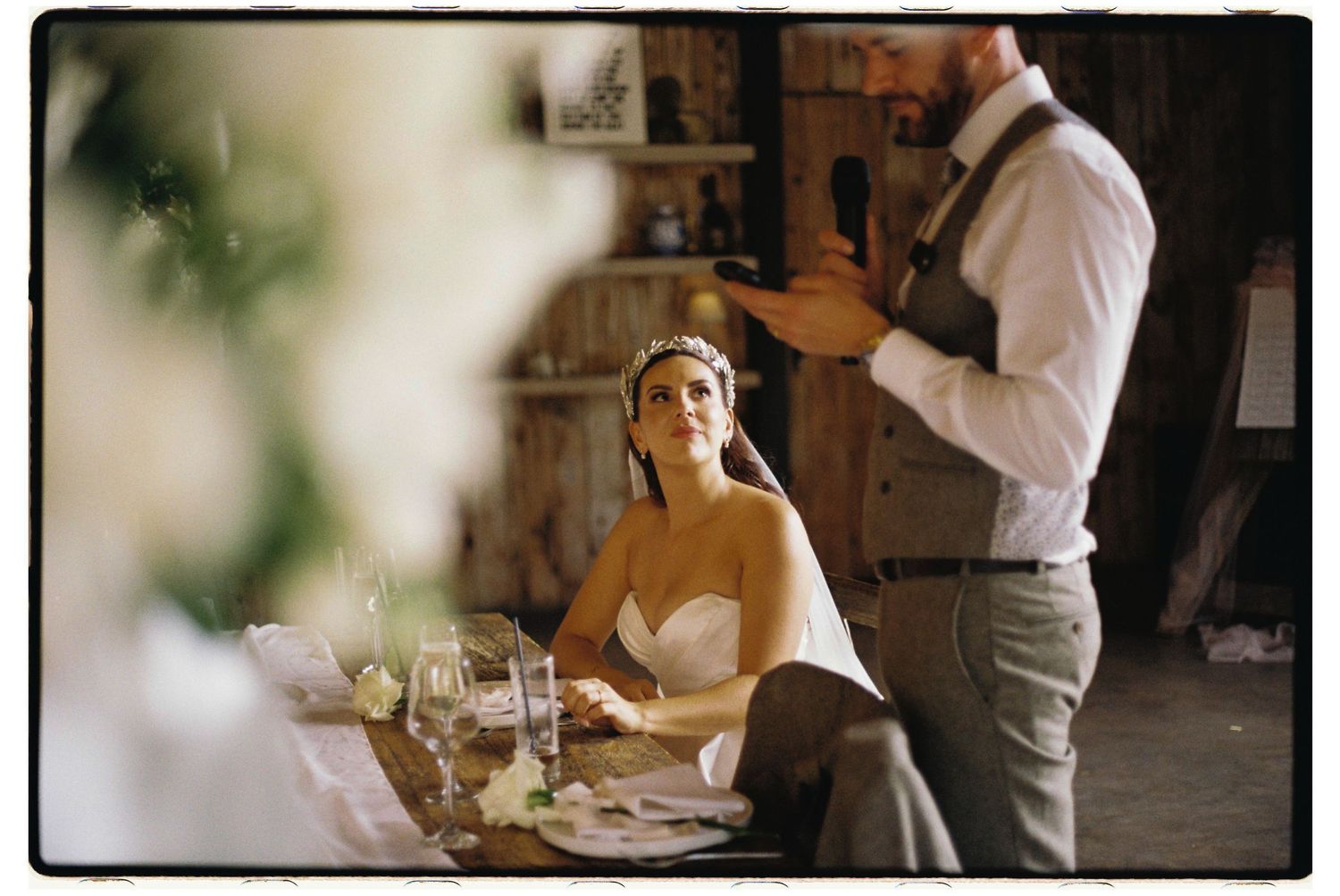 A couple at a rustic restaurant setting during what appears to be a wedding celebration with wine glasses on the table.