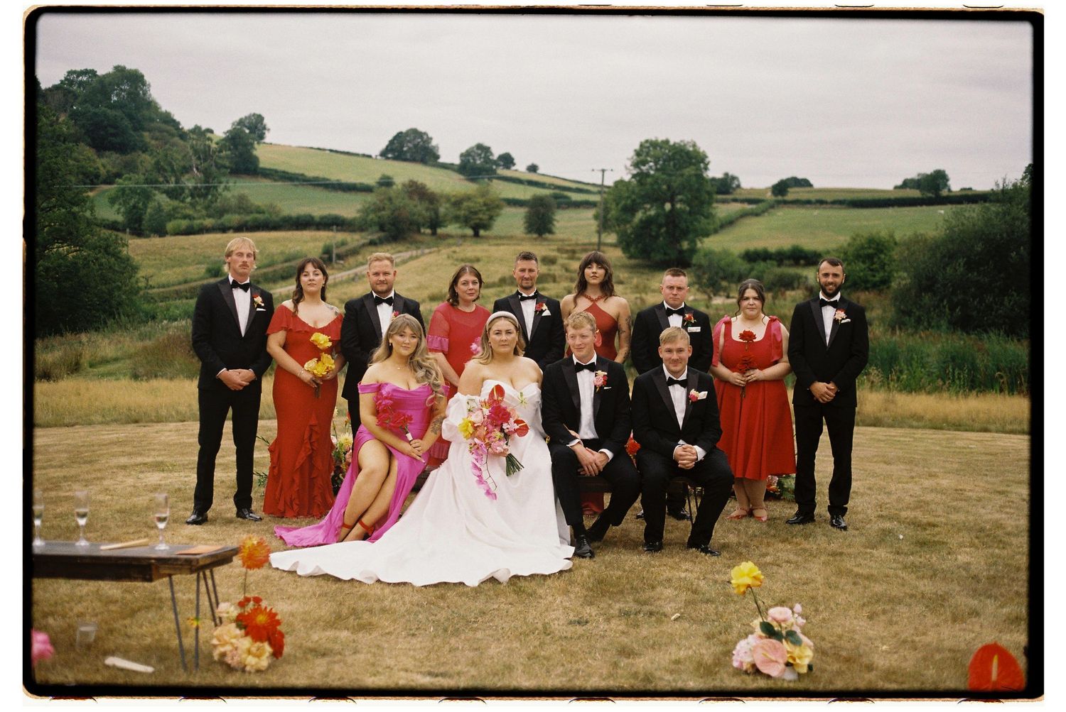 Wedding party in formal attire poses together outdoors against a backdrop of rolling countryside hills.