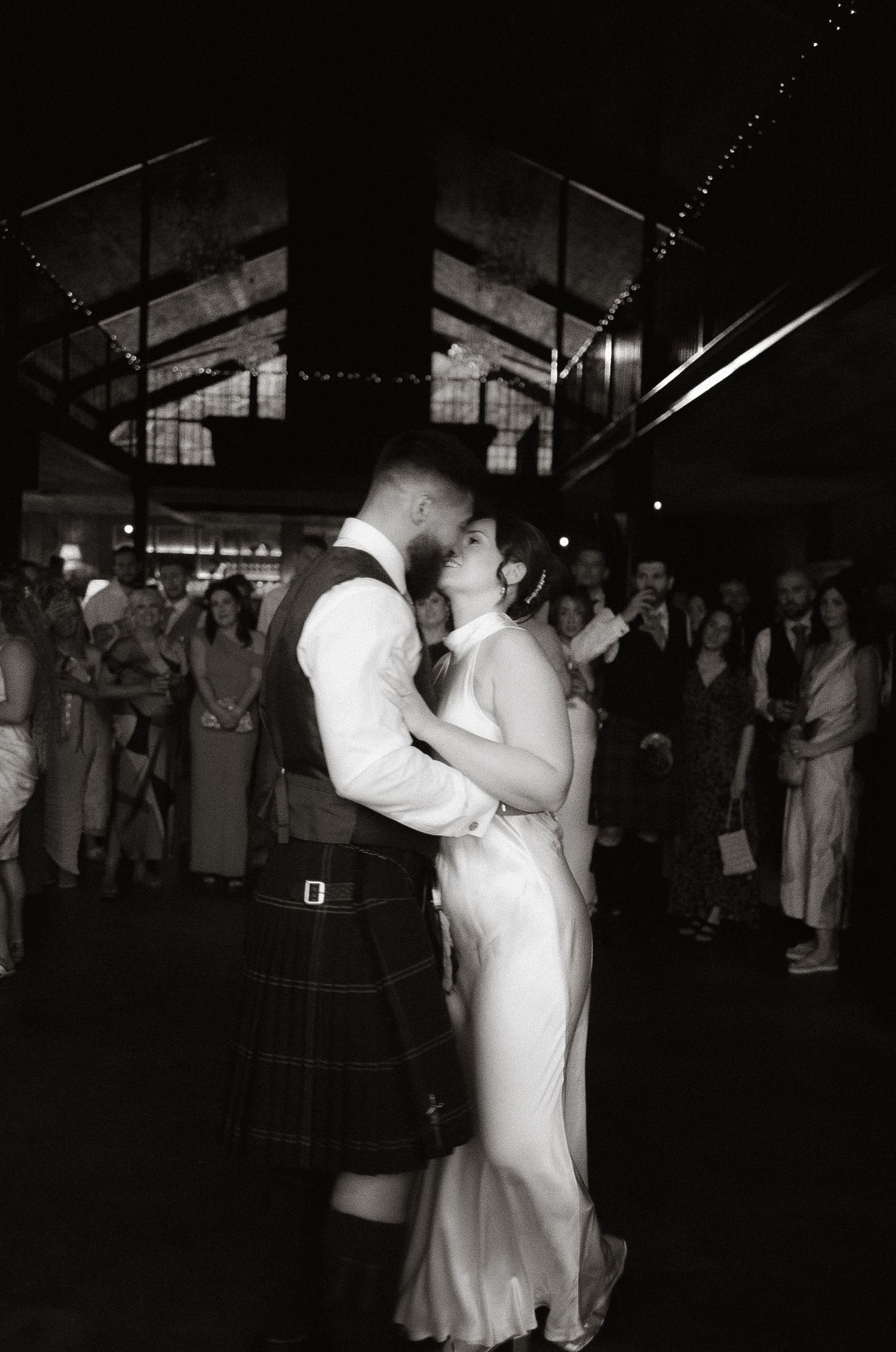 A couple shares their first dance at a wedding reception, with the groom wearing a traditional Scottish kilt under dim lighting.