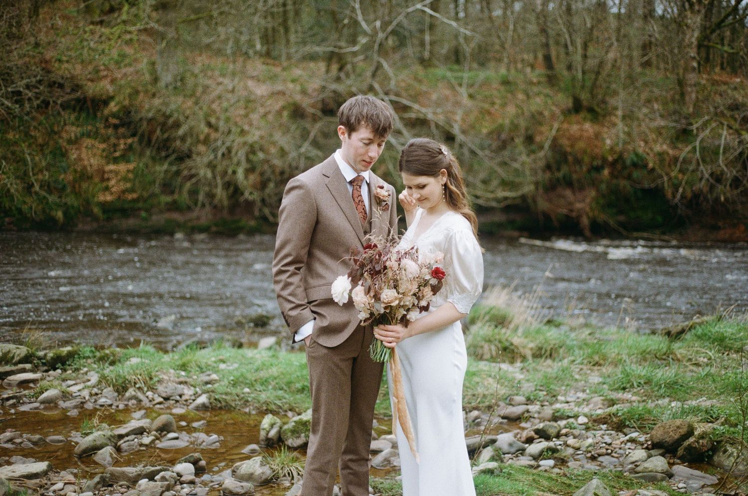 A romantic wedding couple stands beside a river in autumn, with the bride holding a rustic bouquet.