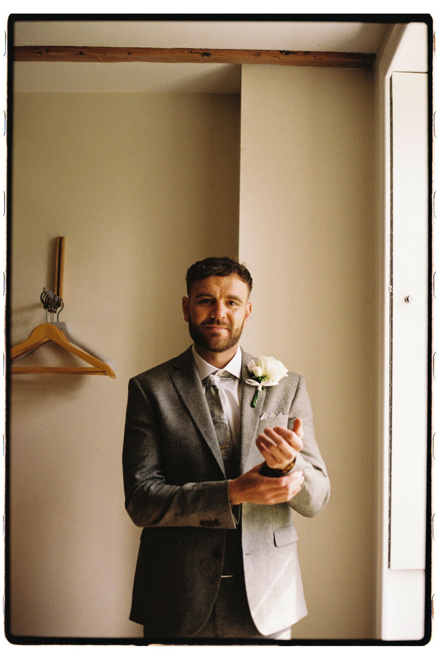 A groom in a grey suit with a boutonniere stands in front of a wooden hanger against a cream-colored wall.