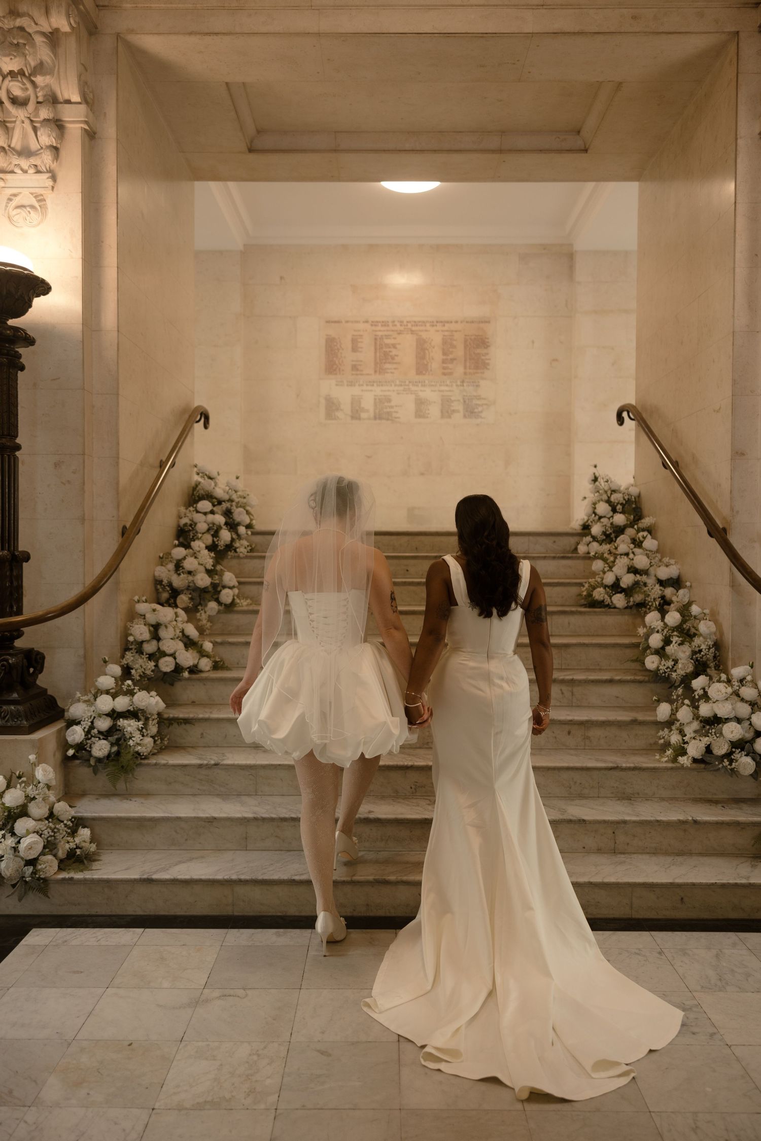 Two brides in white gowns hold hands ascending flower-adorned marble stairs in an elegant wedding venue.