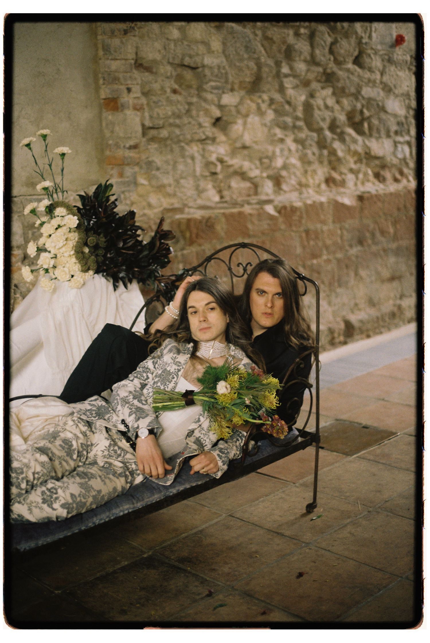 A couple reclines together on a vintage daybed against a rustic stone wall with floral arrangements.
