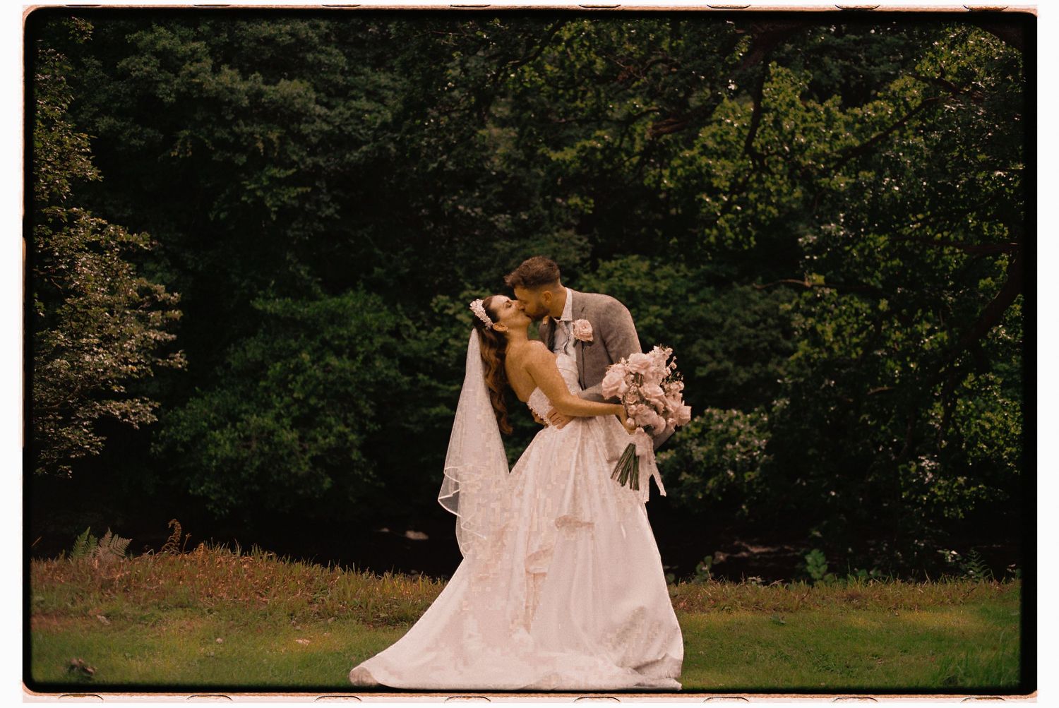 A romantic wedding photograph of a couple sharing a kiss while standing in a lush outdoor garden setting.