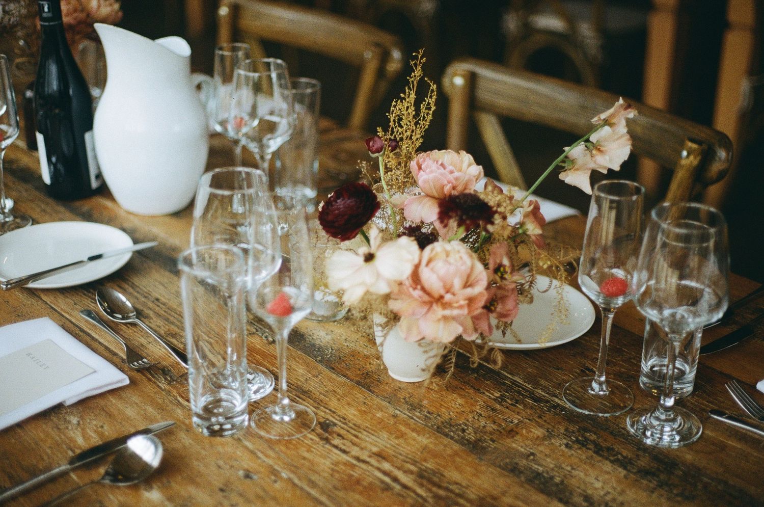 Rustic wedding table setting with peach floral centerpiece, wine glasses and white ceramic pitcher on wooden table.