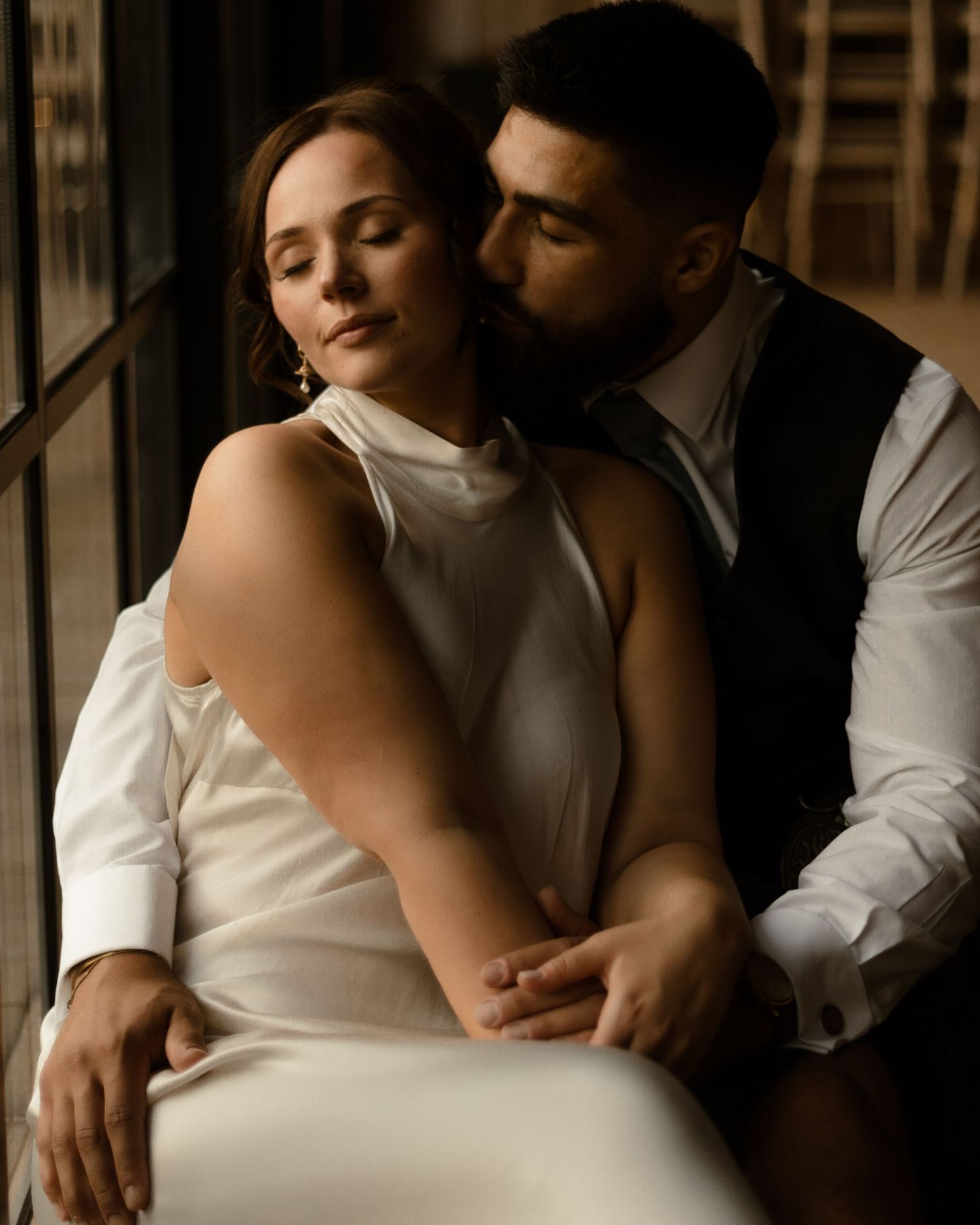 Intimate couple in elegant white attire share romantic moment in moody lighting against window.