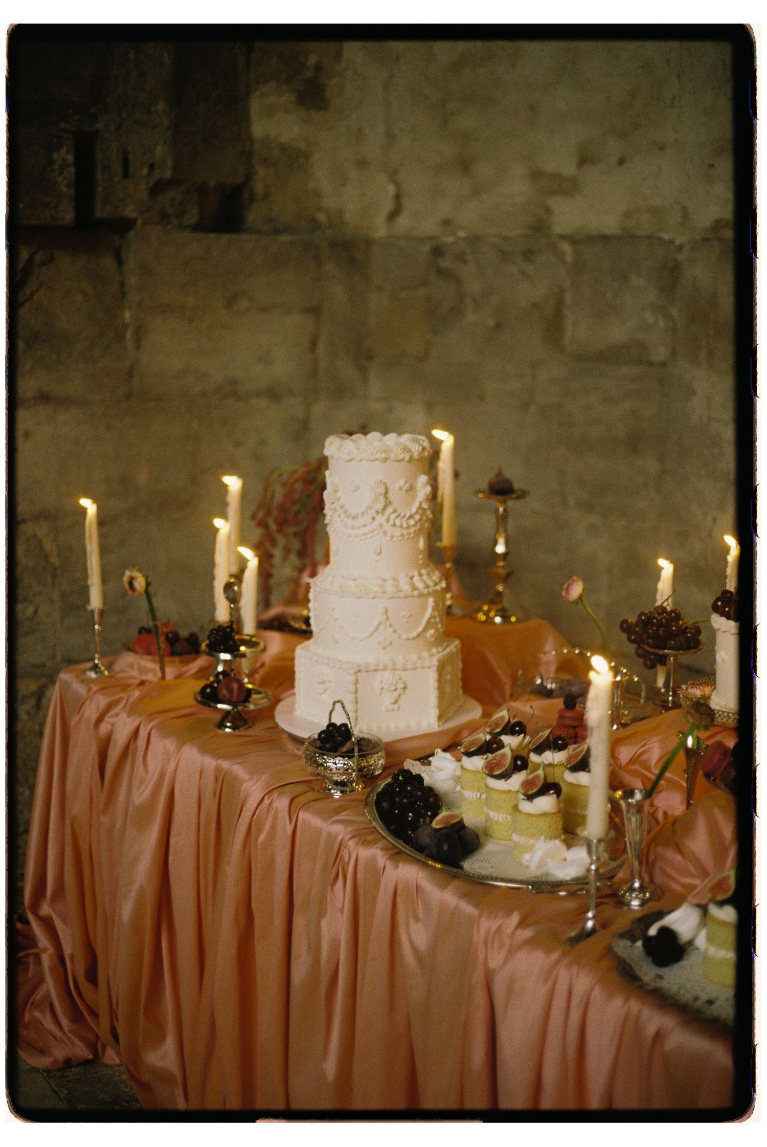 A wedding dessert table decorated with a white tiered cake, candles, and pink tablecloth at an elegant reception.
