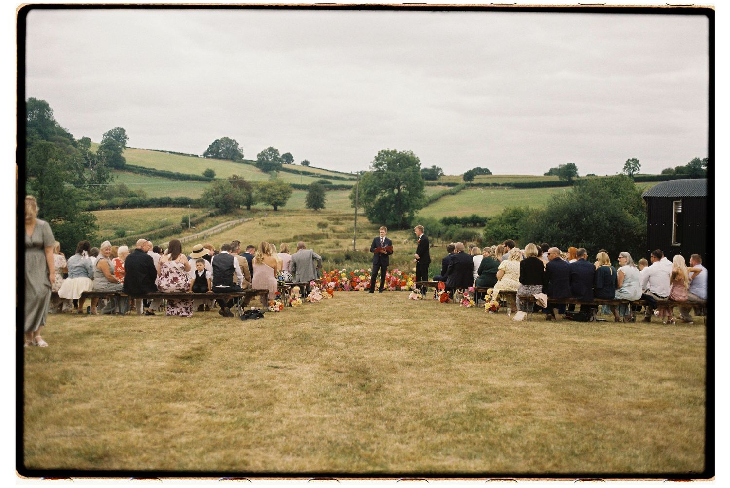 People gathered for an outdoor event on a grassy hillside in rural countryside during summer.