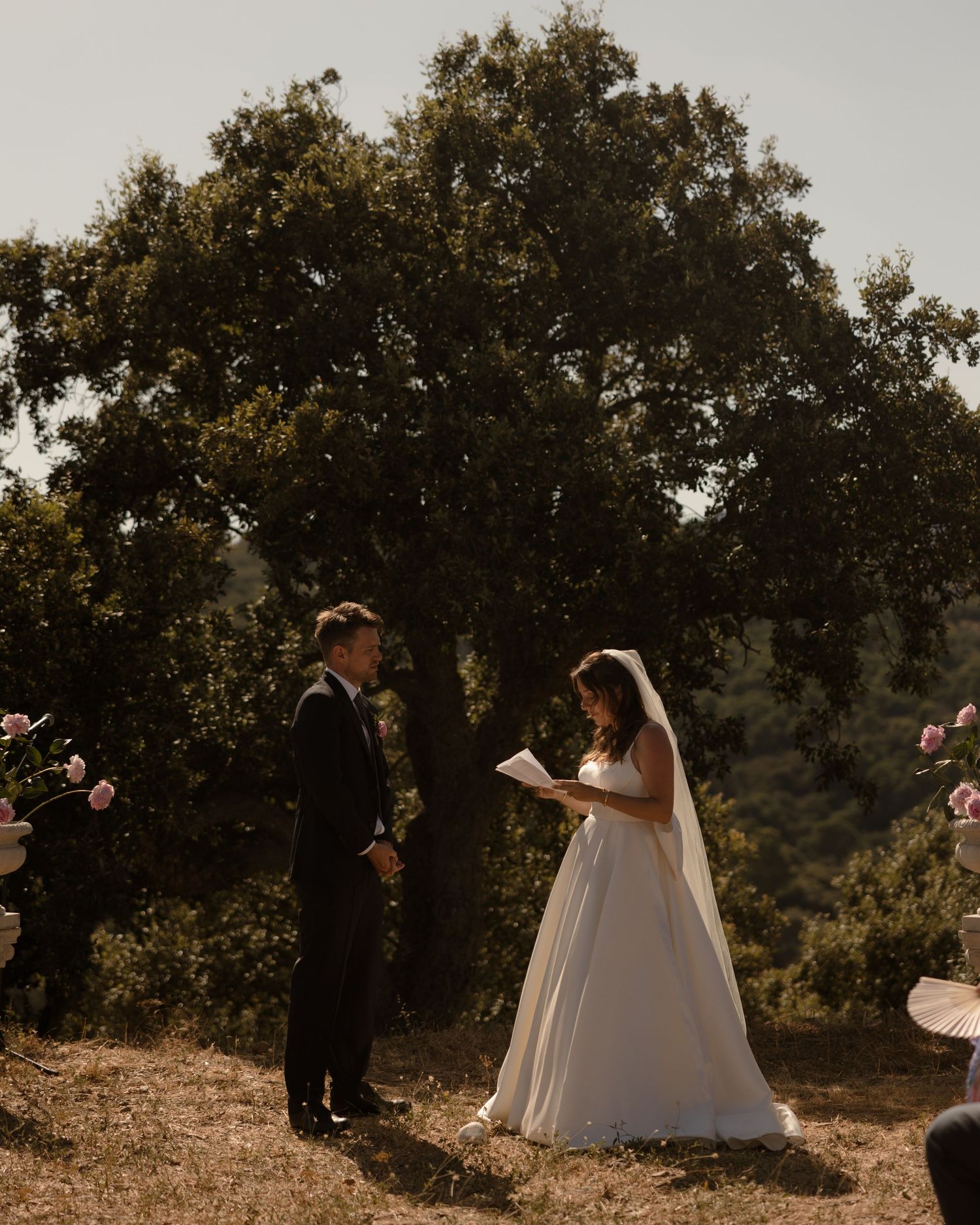 A couple exchanges vows during their outdoor wedding ceremony under a large oak tree on a sunny day.