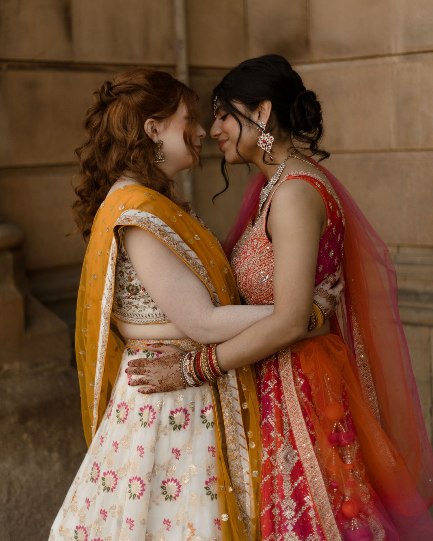 Two people in traditional Indian bridal attire embrace in soft lighting against a stone wall backdrop.