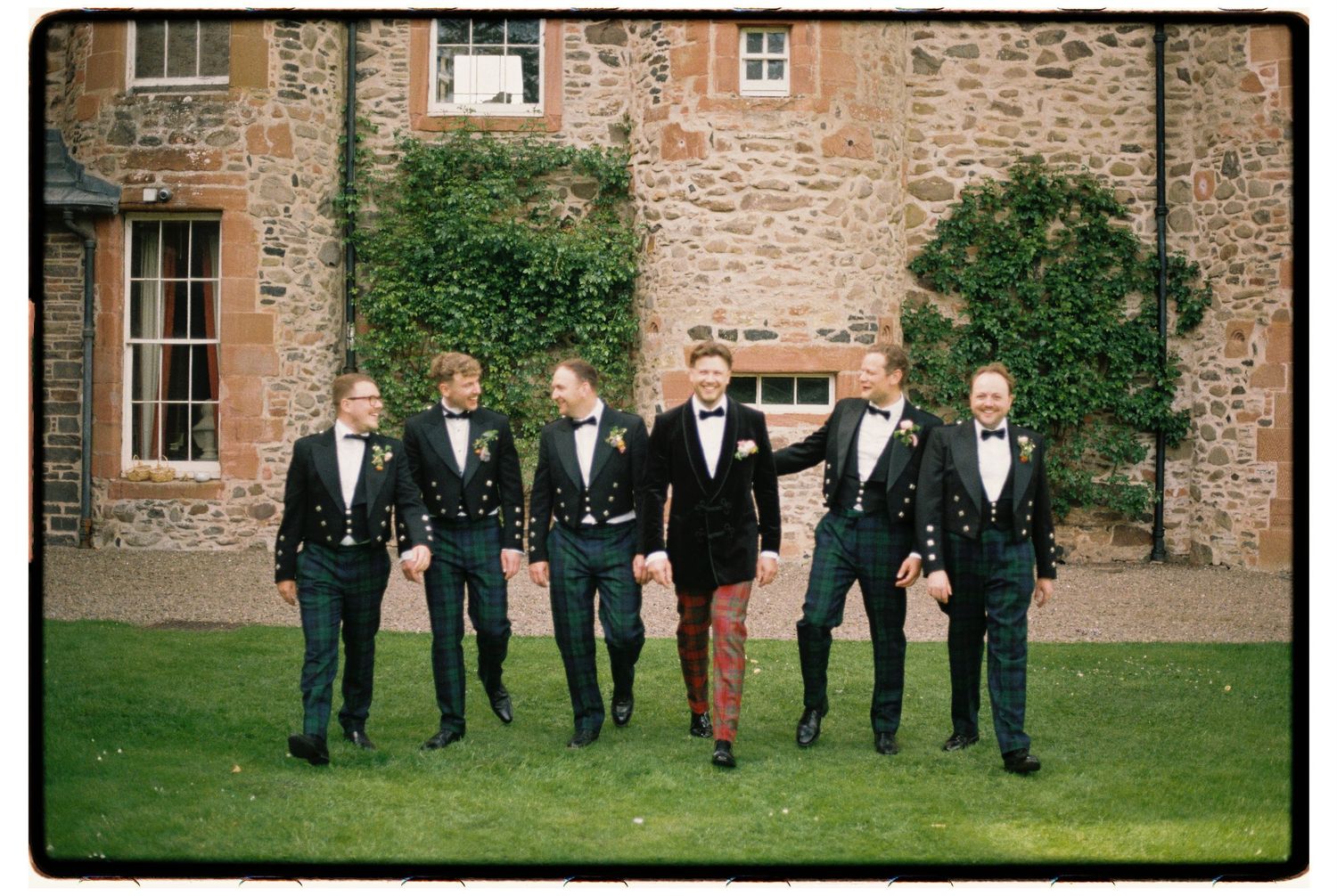 Groom and groomsmen in black tuxedos walk together on lawn outside stone manor house wedding venue.