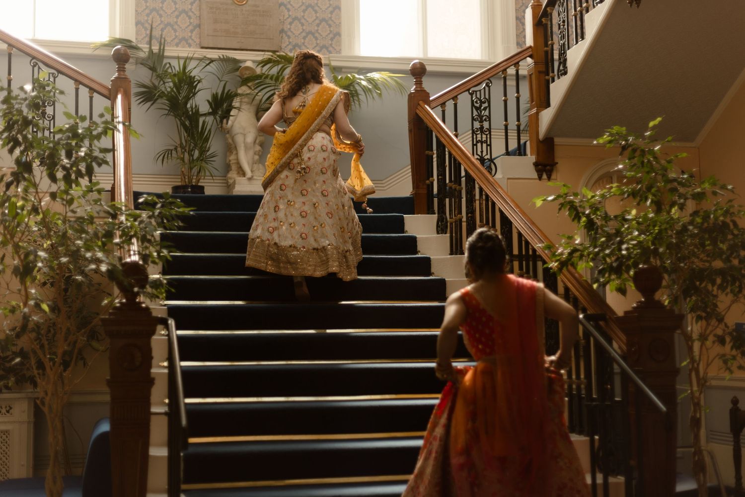 Two individuals in flowing traditional dresses descend an elegant staircase adorned with potted plants in warm lighting.