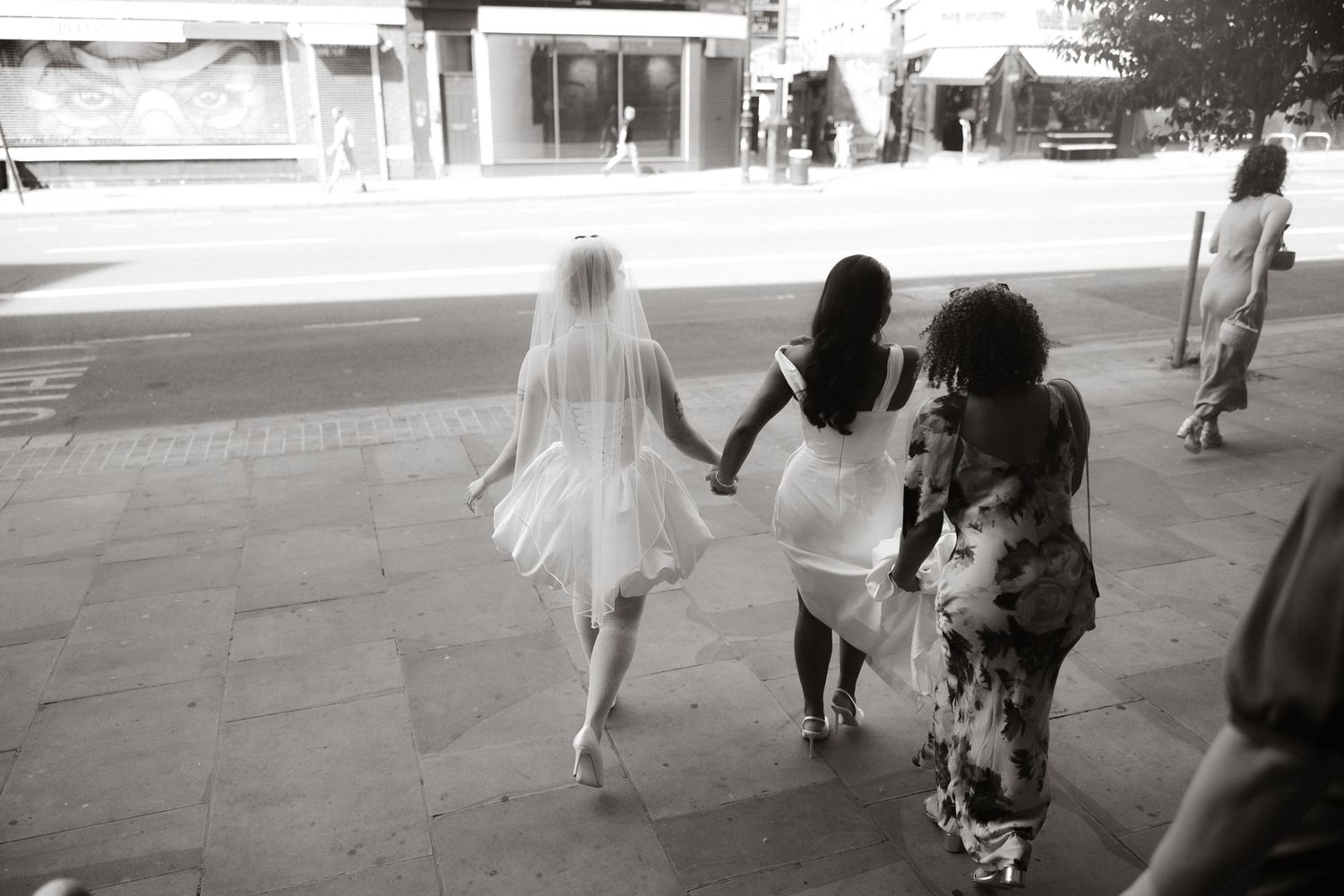 Black and white photo of a bride in veil and two women walking together on a city sidewalk.