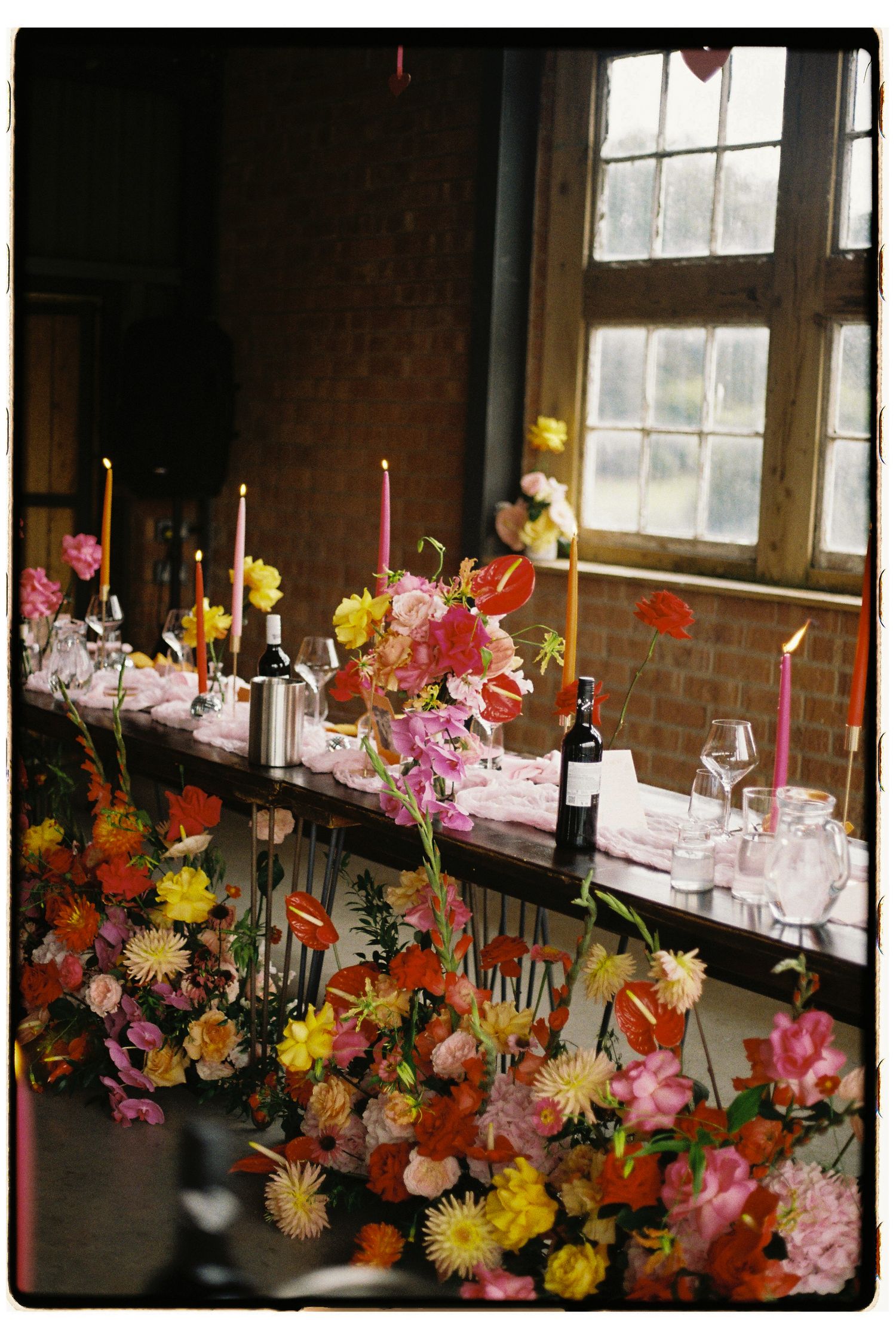Floral table arrangement with wine bottles and candles set near a rustic window, featuring colorful blooms and place settings.