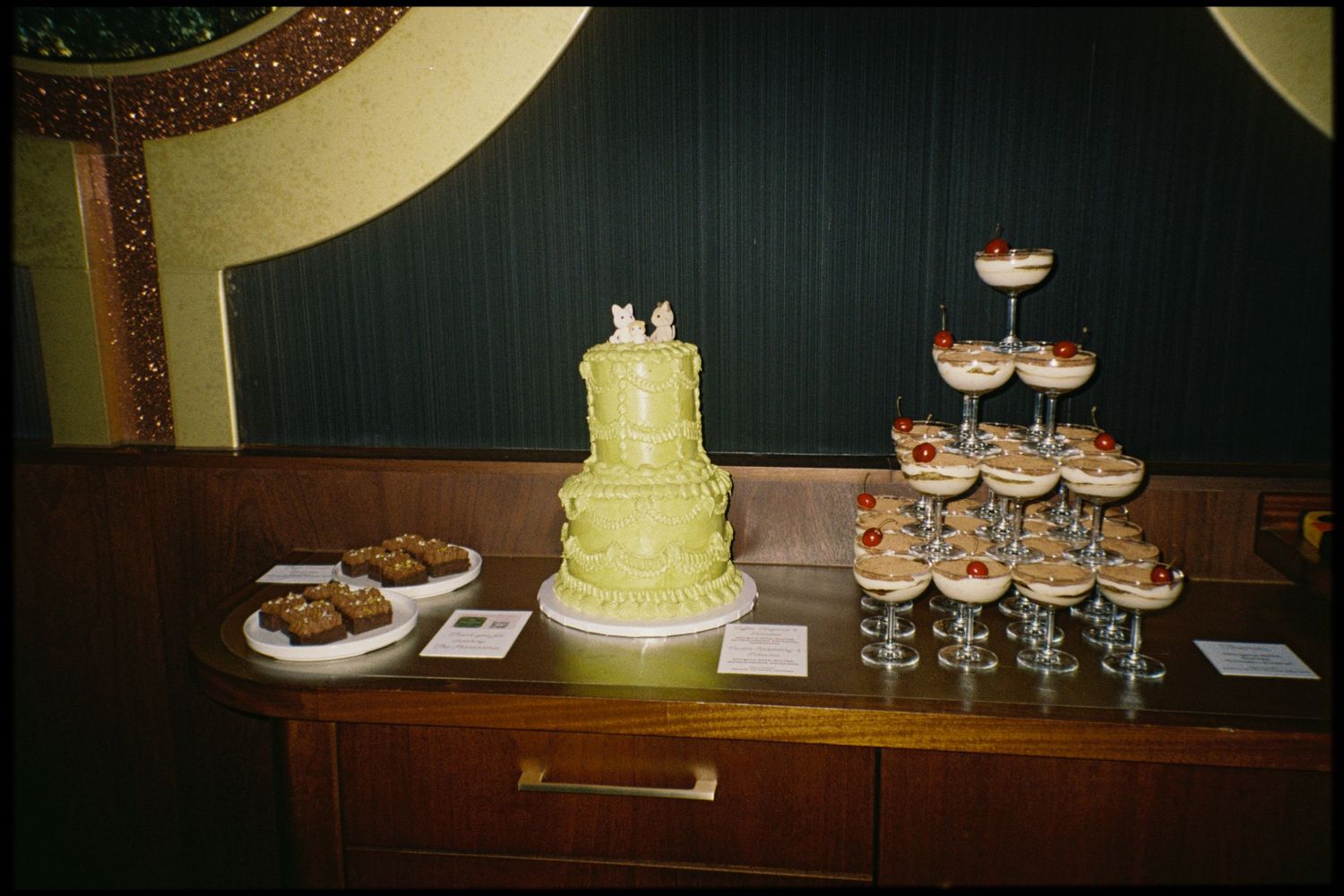 A yellow wedding cake and dessert table display with cupcakes and glasses arranged at an elegant reception.