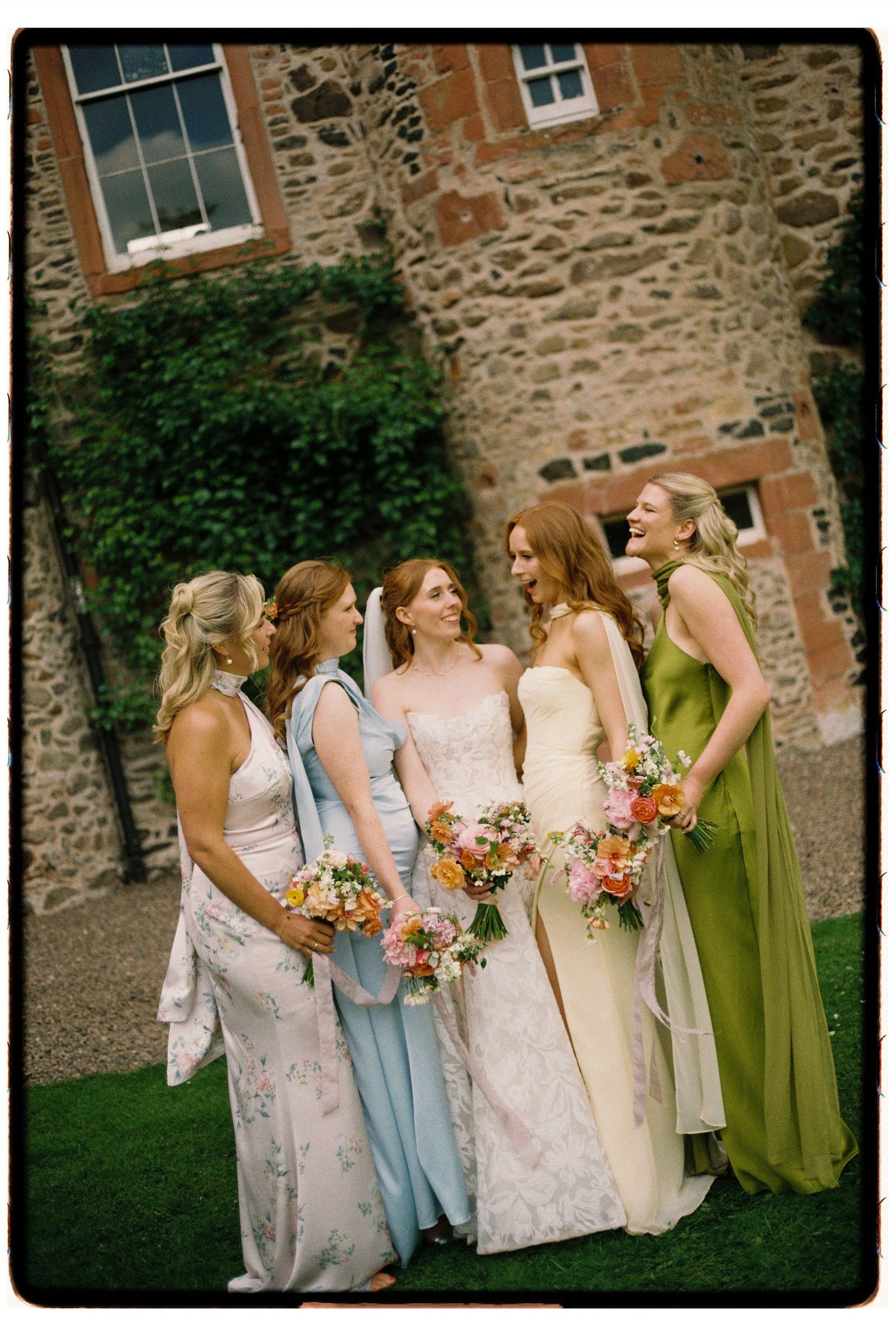 Bride and four bridesmaids in colorful gowns holding bouquets, laughing together outside a stone building.