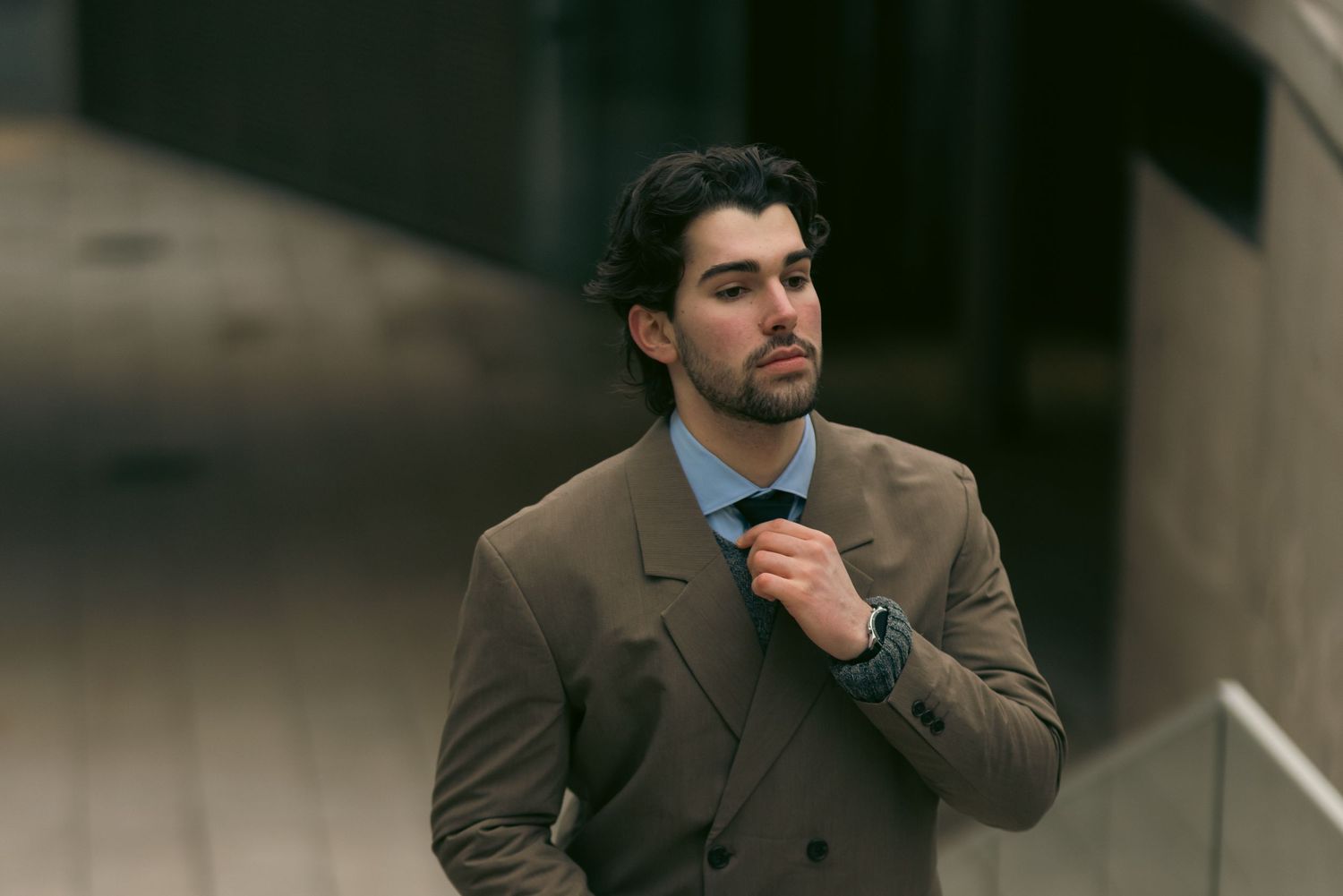 Stylish man in brown double-breasted suit adjusting tie outdoors near modern building.