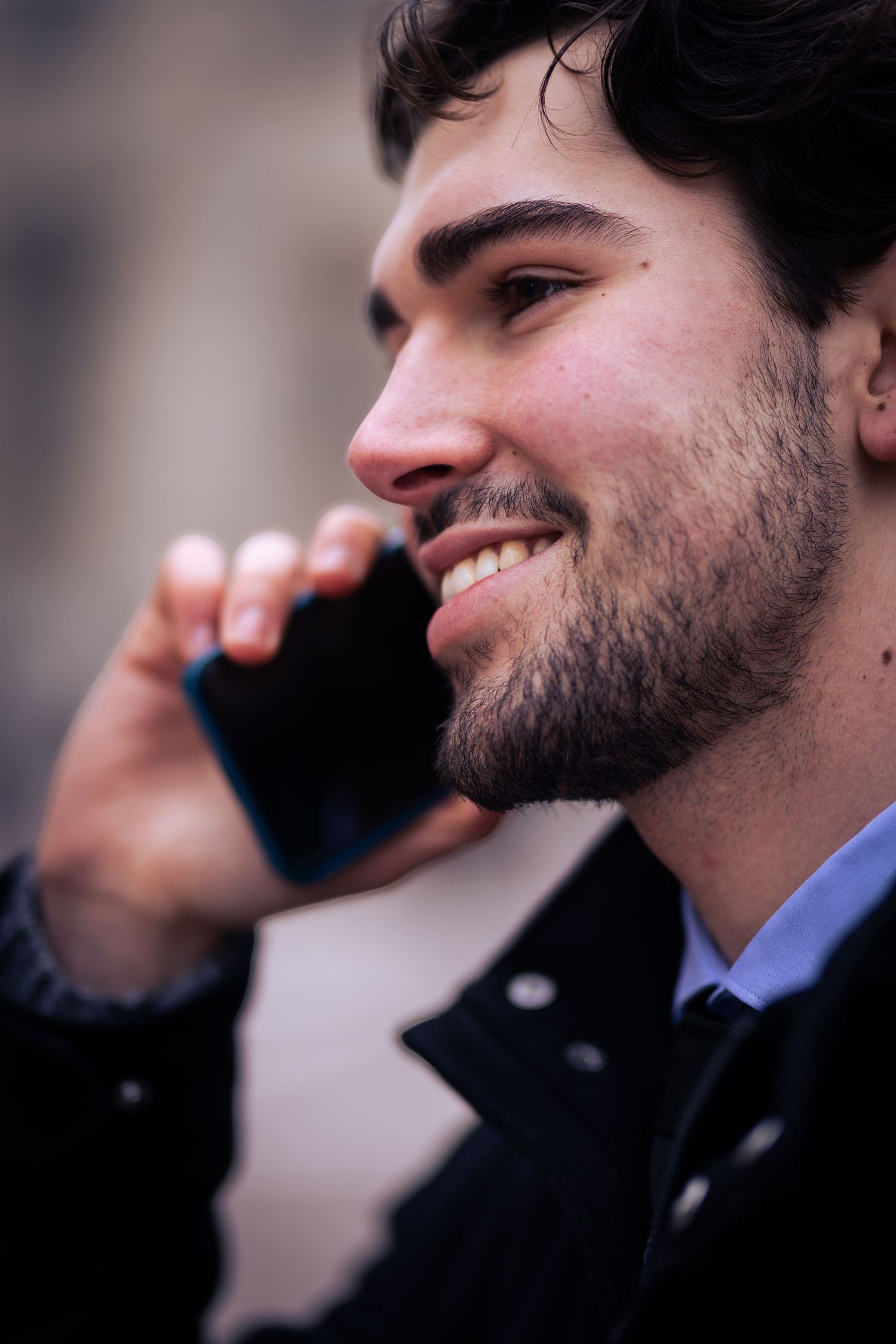 Smiling young bearded man in black jacket talking on smartphone outdoors.