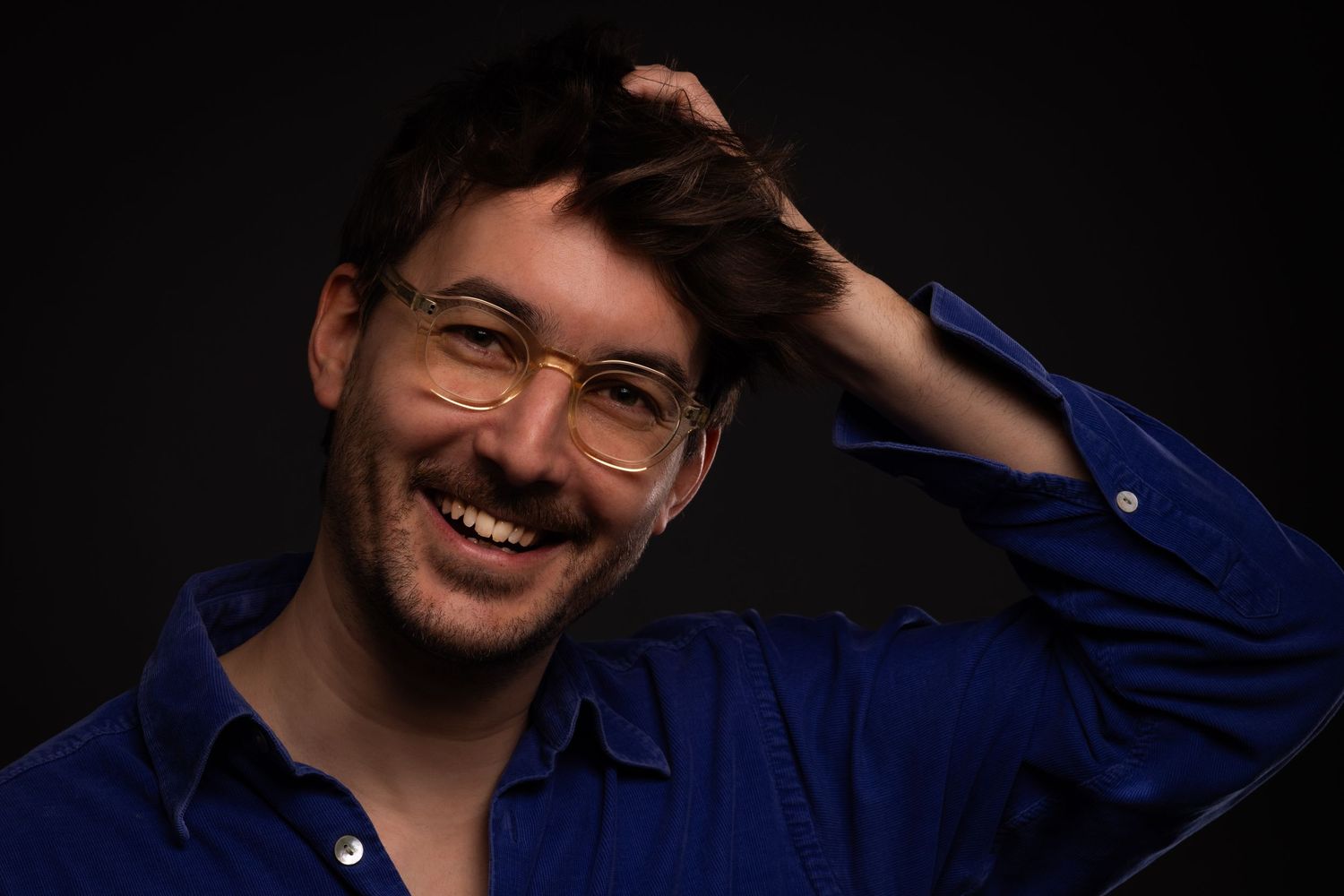 Cheerful young man with glasses and blue shirt laughing while running hand through hair on dark background.