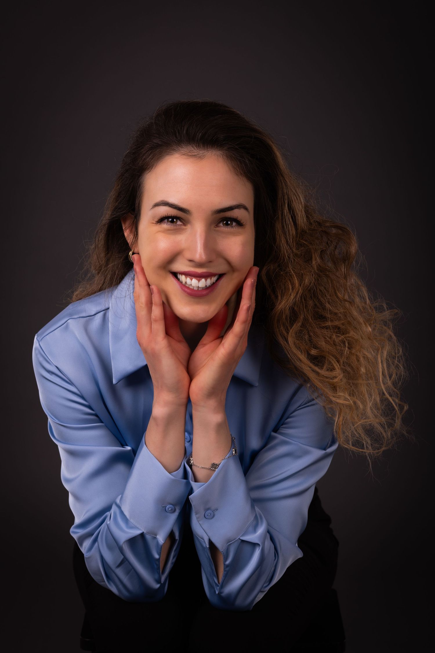 Smiling young woman with curly hair in blue shirt holding face with both hands against dark background.