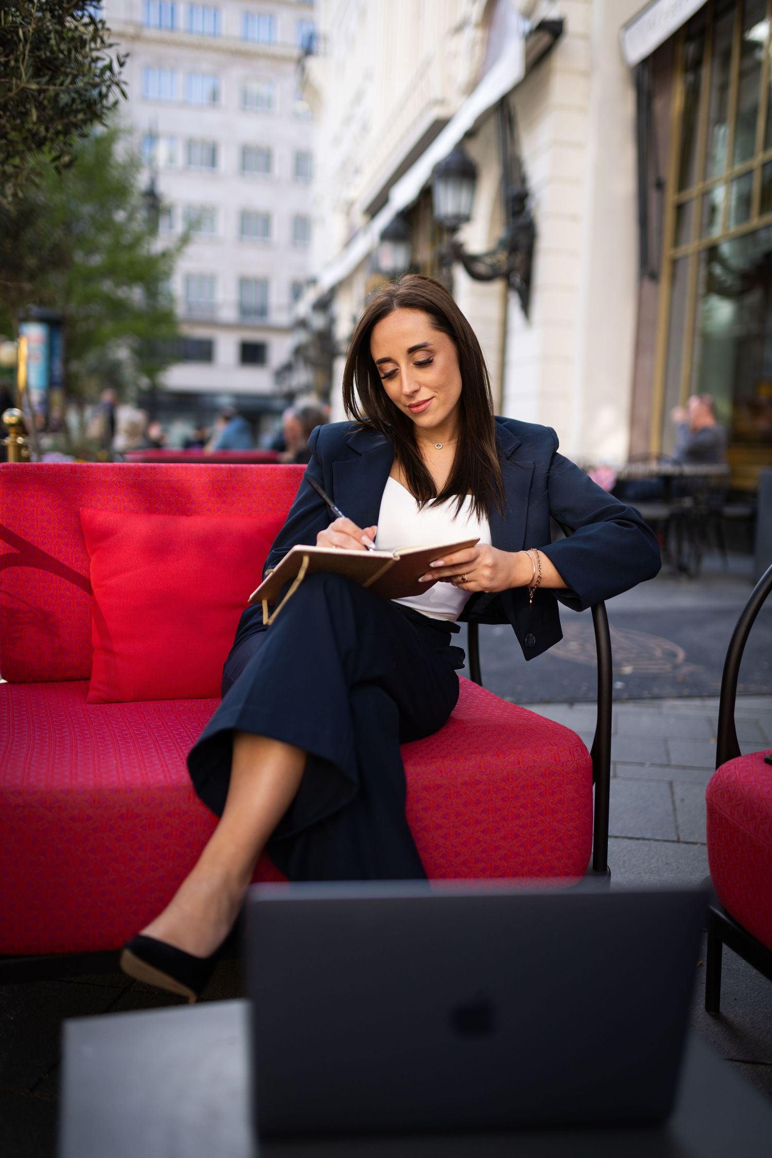 Professional woman in navy suit writing in notebook while sitting on red outdoor sofa.