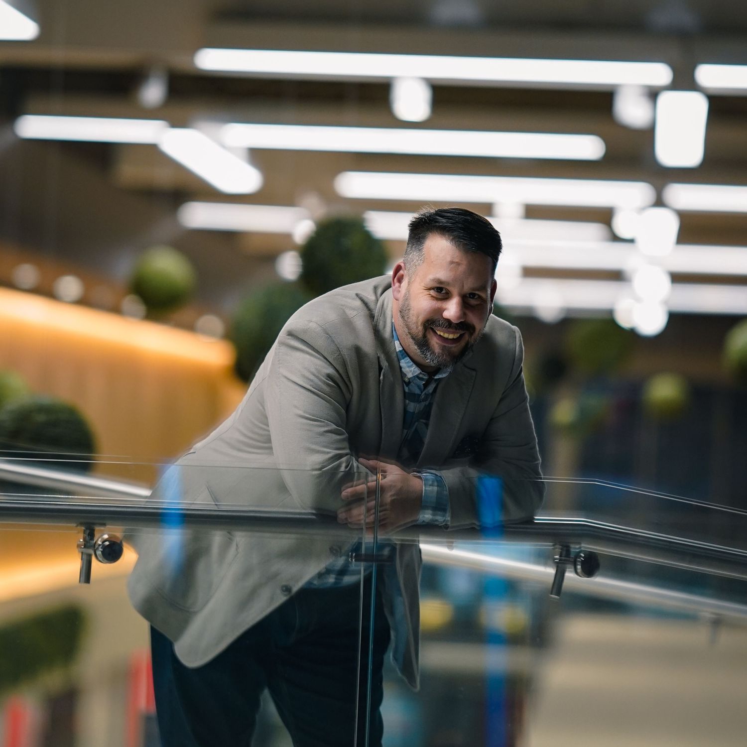 Smiling bearded businessman in grey blazer leaning on glass railing in a modern office building interior.