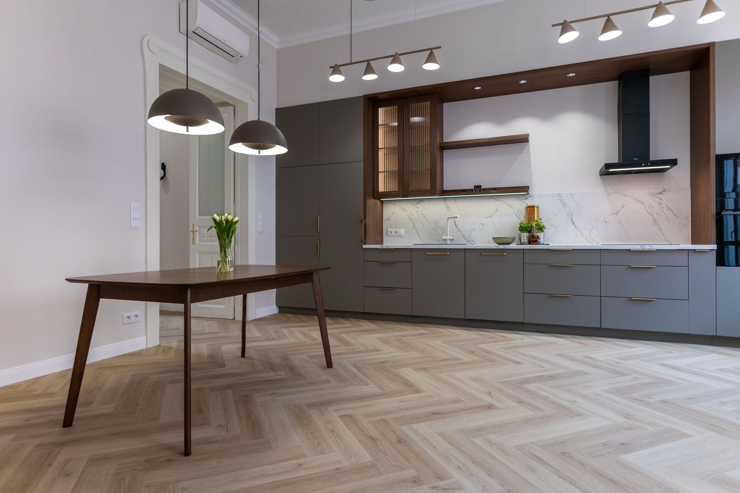 Modern kitchen with grey cabinets, marble backsplash, herringbone wood floors, and pendant lights over dining table.