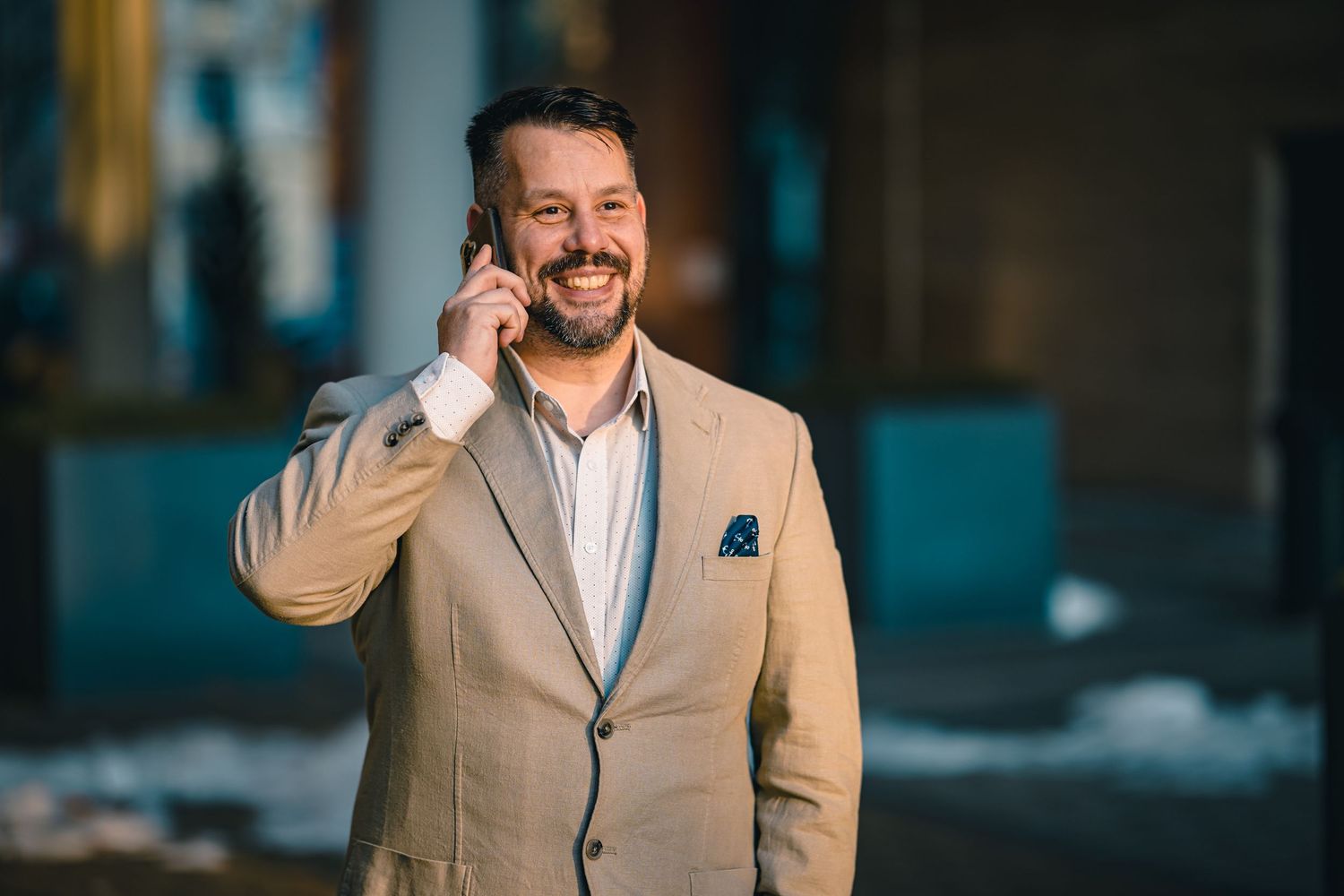 Happy businessman in beige blazer smiling while talking on phone outdoors.