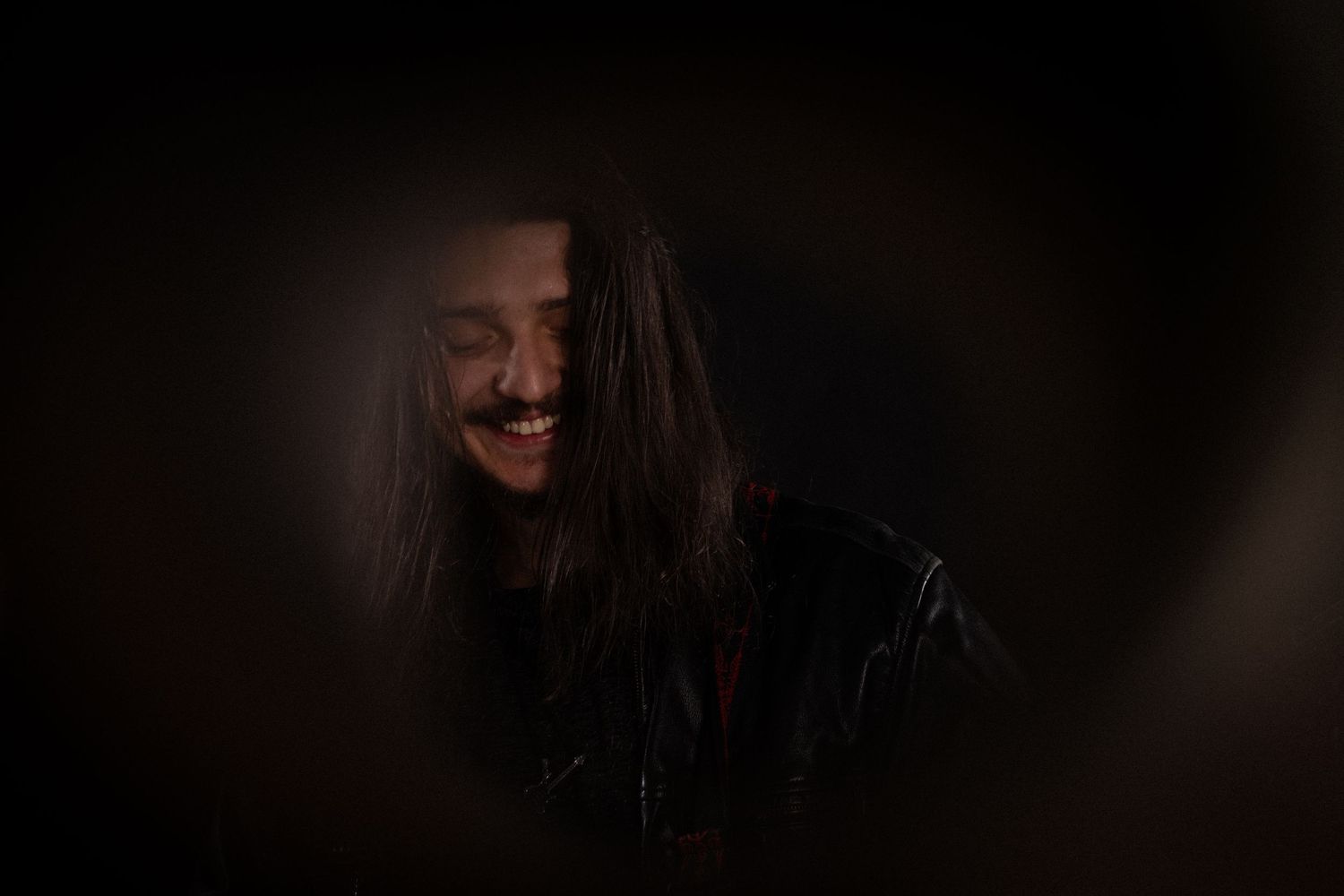 Young person with long hair laughing in dramatic dark lighting with leather jacket.