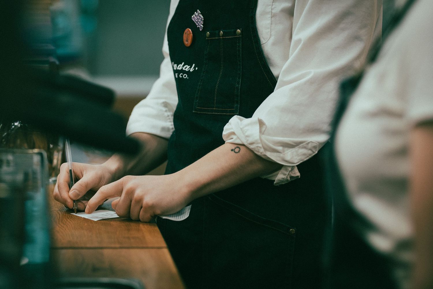 Barista in denim apron writing on notepad at wooden counter in cafe.