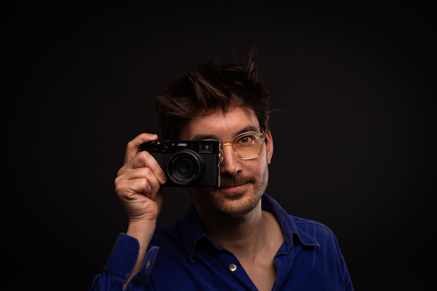 Young man with glasses holding a compact camera up to his eye against a dark studio background.