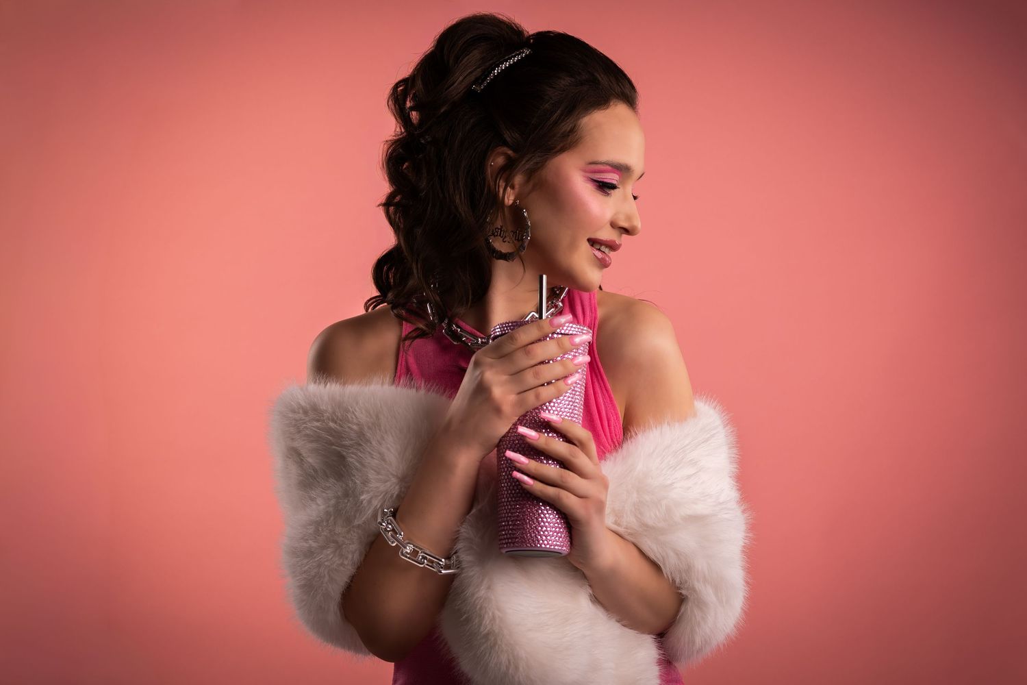 Elegant young woman in pink dress with white fur stole holding a drink against a pink studio background.
