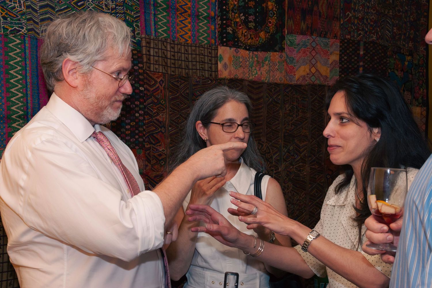 Three people engaged in conversation at a social gathering with colorful tapestries in the background.