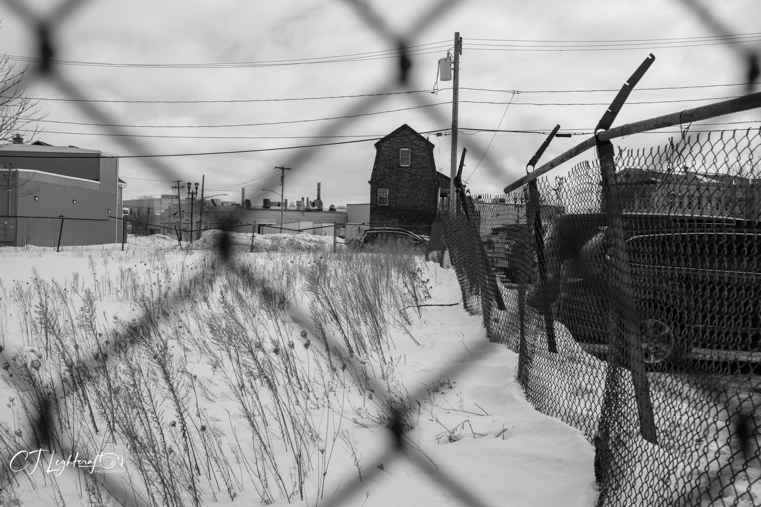 Black and white photo of a snowy urban landscape viewed through a chain-link fence with buildings in background.