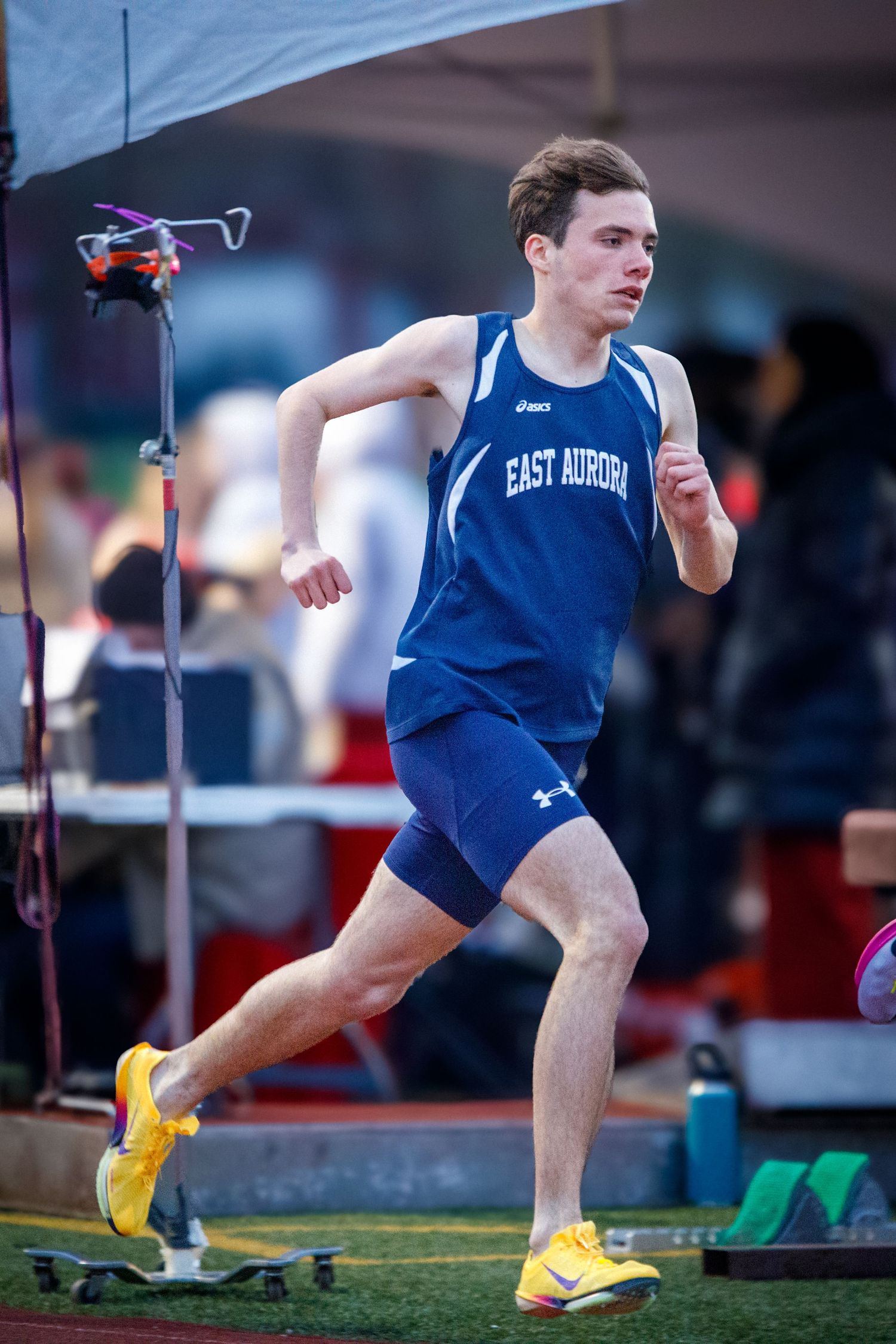 A male East Aurora track athlete in blue uniform runs during an indoor track competition.