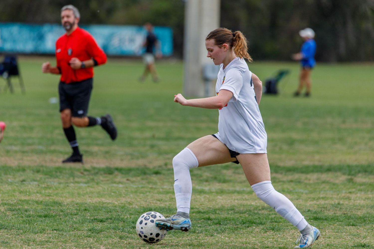 A female soccer player in white kicks a ball on a grass field with a referee running behind her.