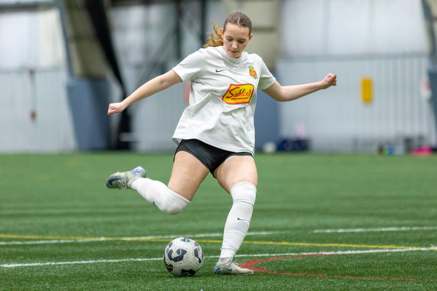 A female soccer player in a white uniform kicks a ball on an indoor turf field.