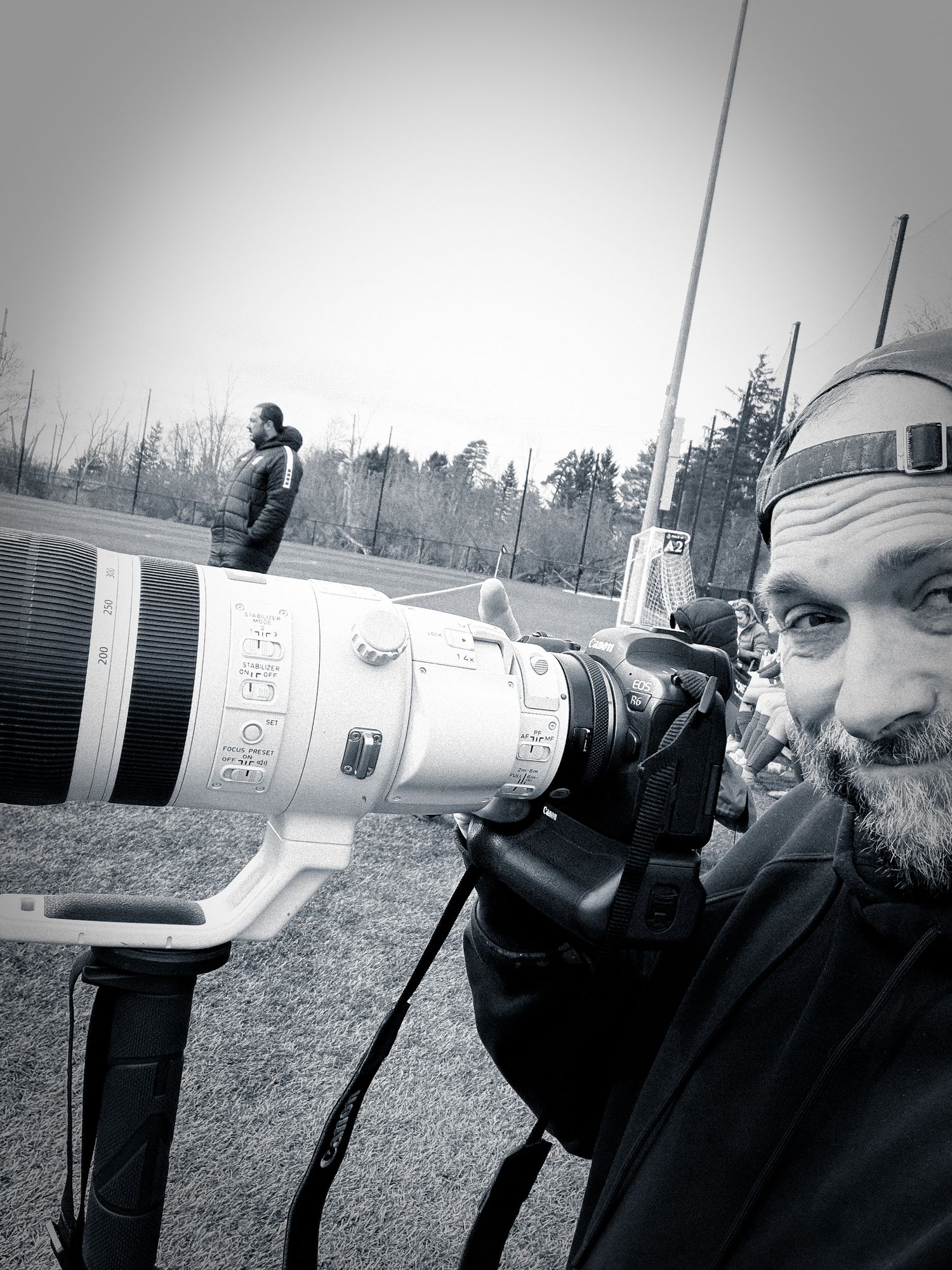 Black and white selfie of male photographer holding professional camera with large telephoto lens outdoors.