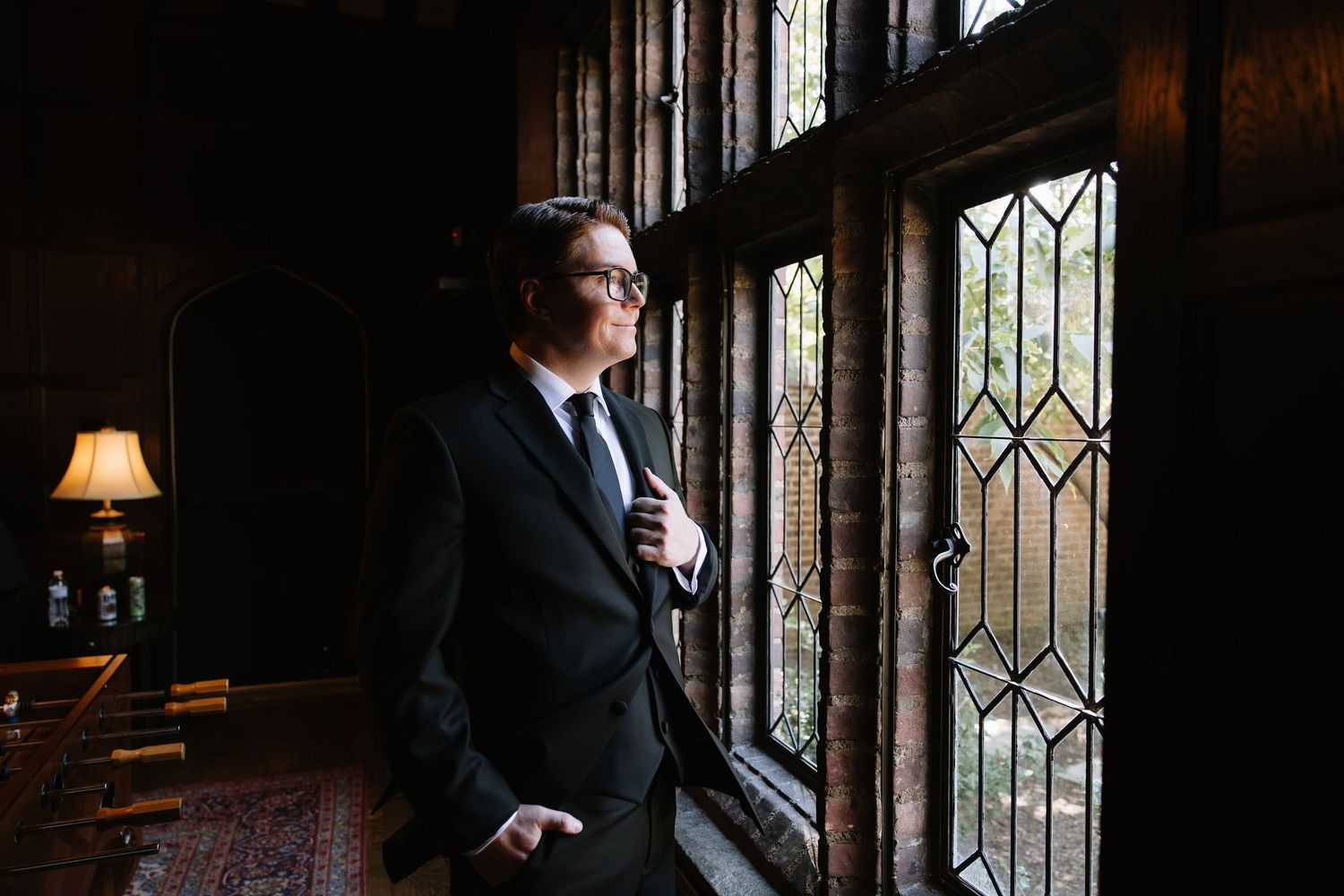 Elegant groom in black suit adjusting tie by ornate leaded glass windows in a dark wood-paneled room.