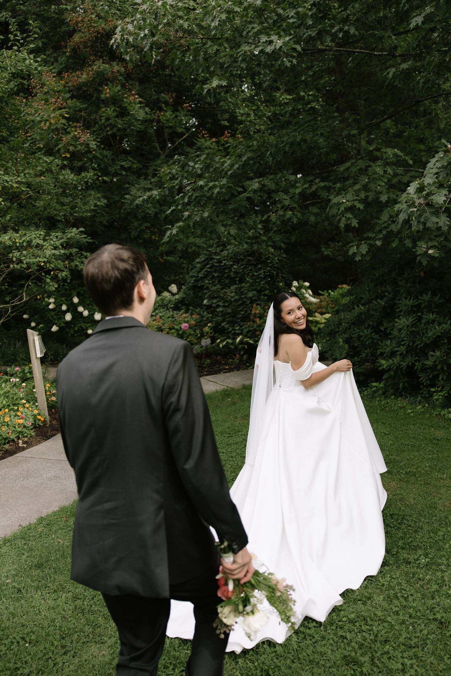 Bride in white gown looks back at groom in black suit holding bouquet in lush garden wedding setting.